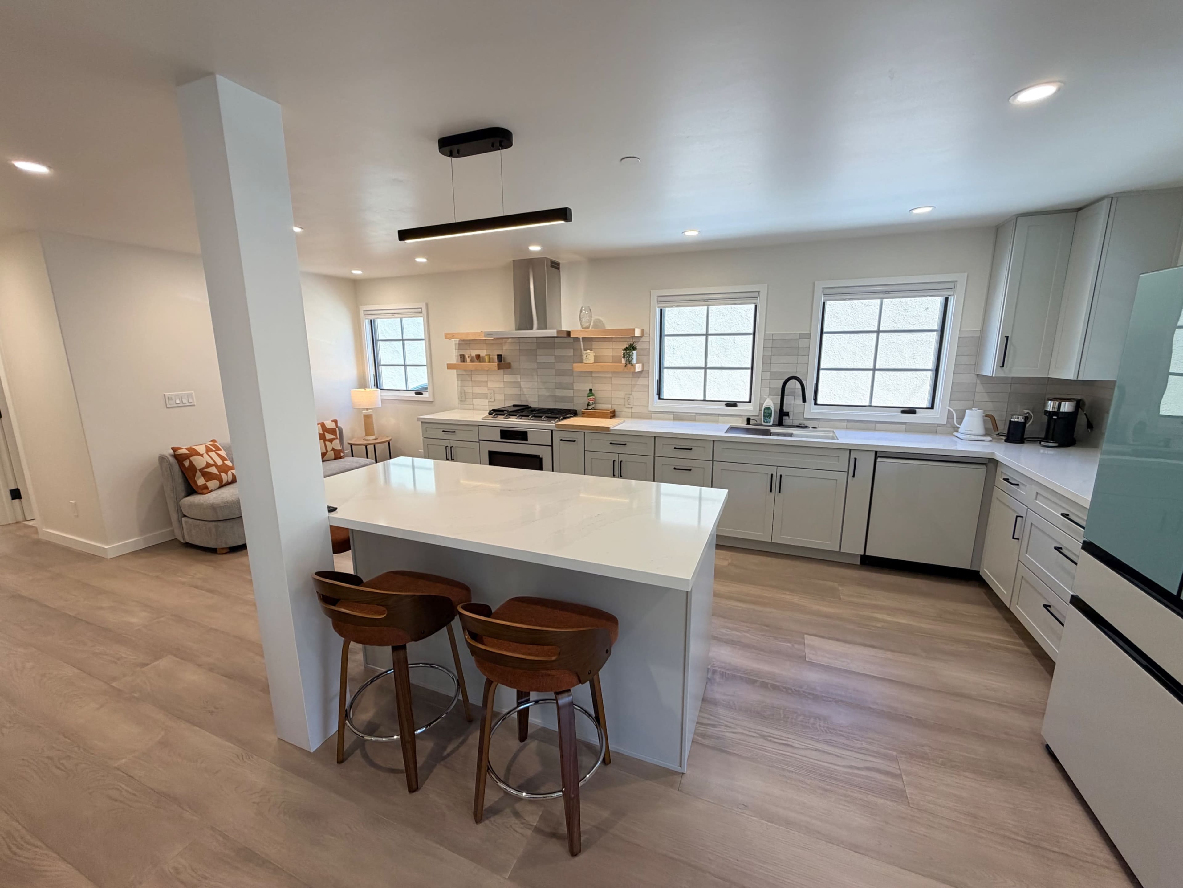 The image shows a modern kitchen featuring a large island with bar stools, white cabinetry, and a mix of appliances, all illuminated by natural light from multiple windows.