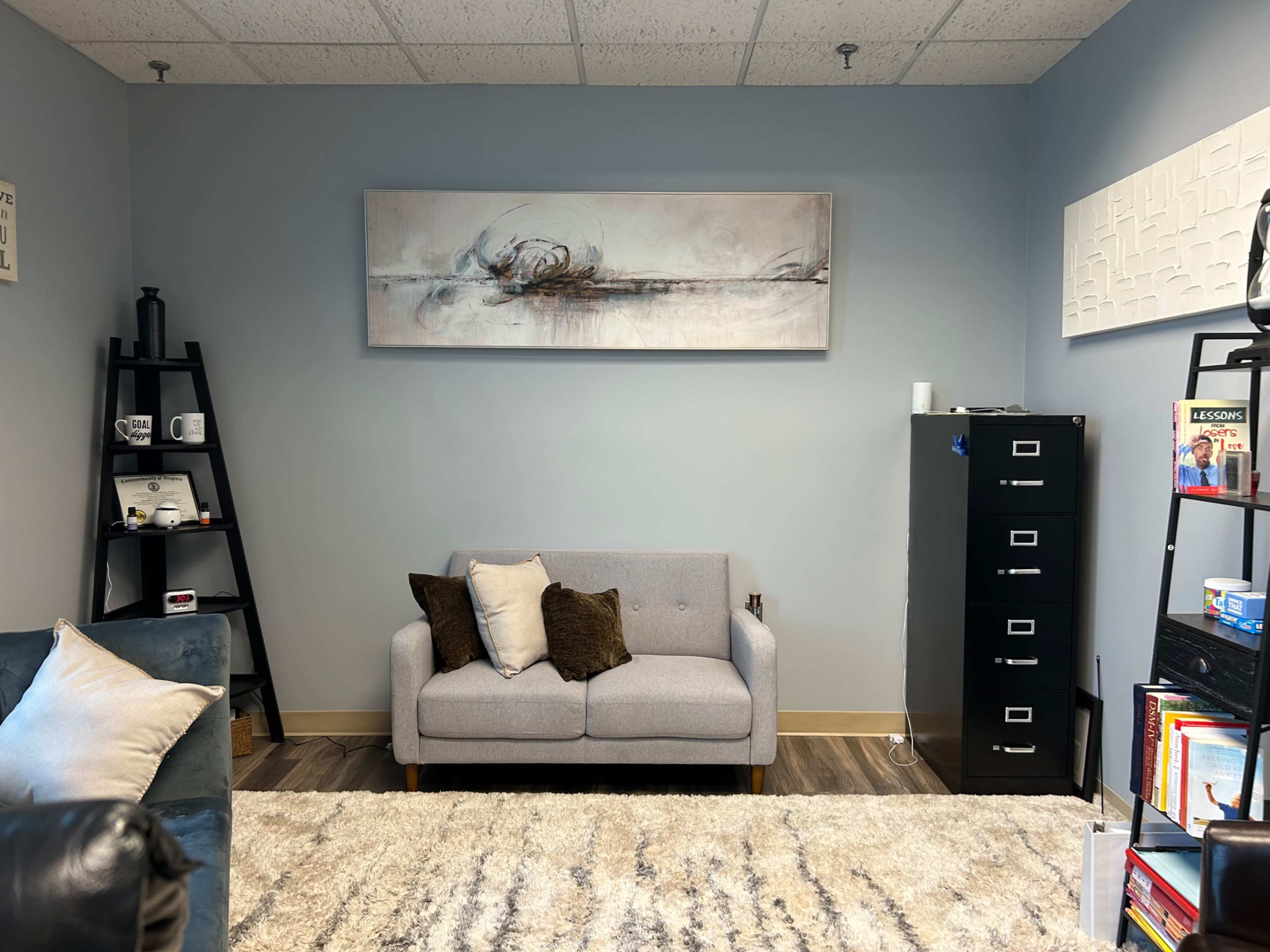 The image shows a modern office lounge area featuring a gray sofa with decorative pillows, a black filing cabinet, bookshelves, and wall art in a softly colored room.