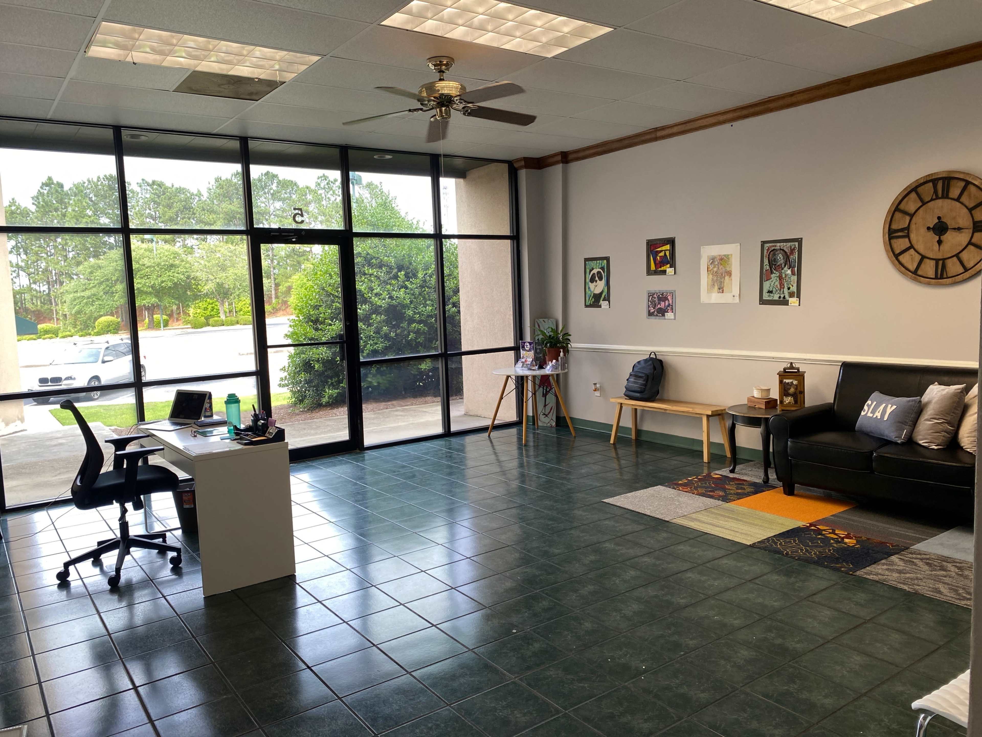 The image shows an empty office space with a black sofa, a small desk, and large windows allowing natural light to enter.