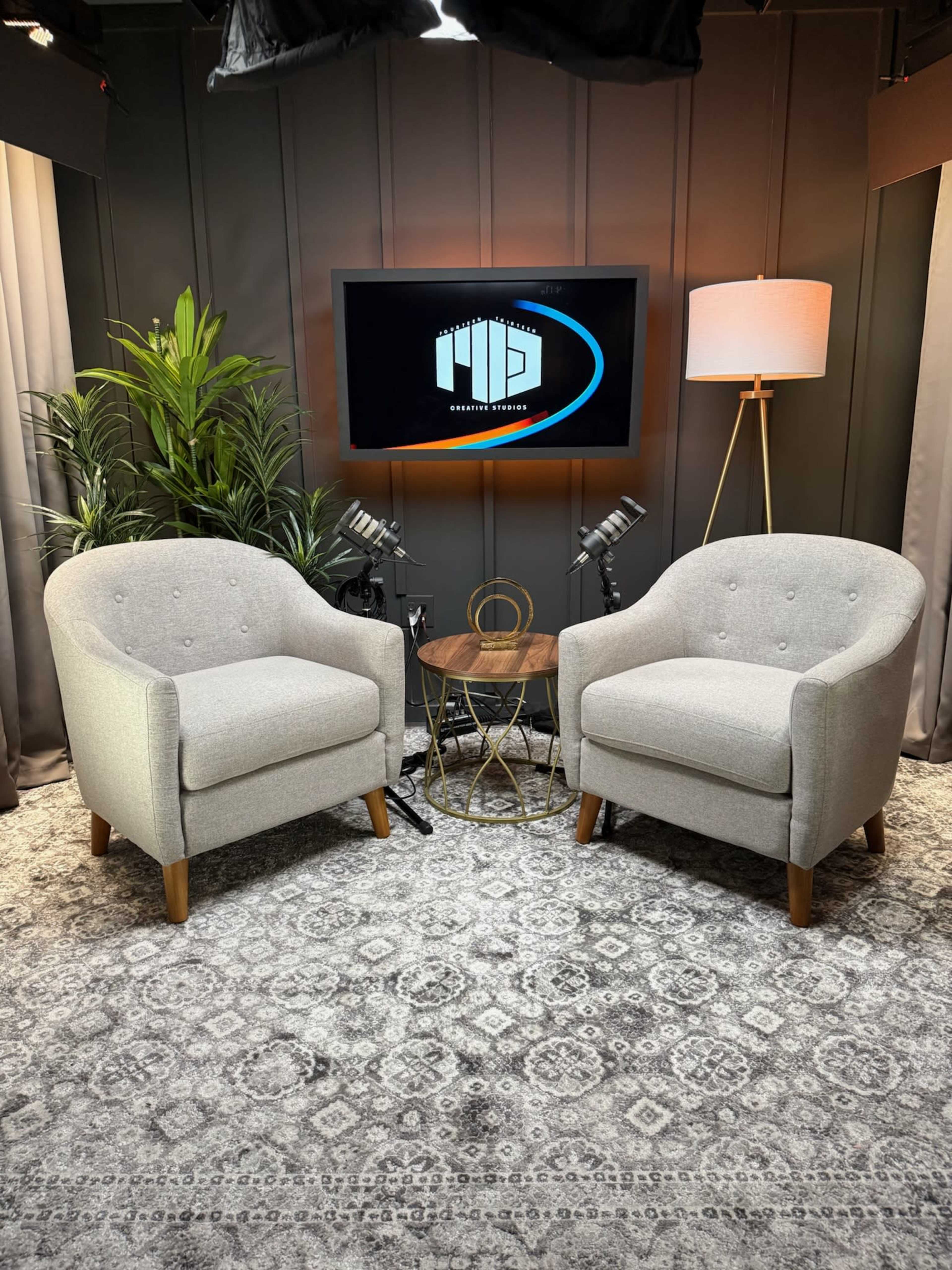 The image shows a modern studio setting featuring two light gray armchairs, a small round table, and decorative plants, with a television displaying a logo in the background.