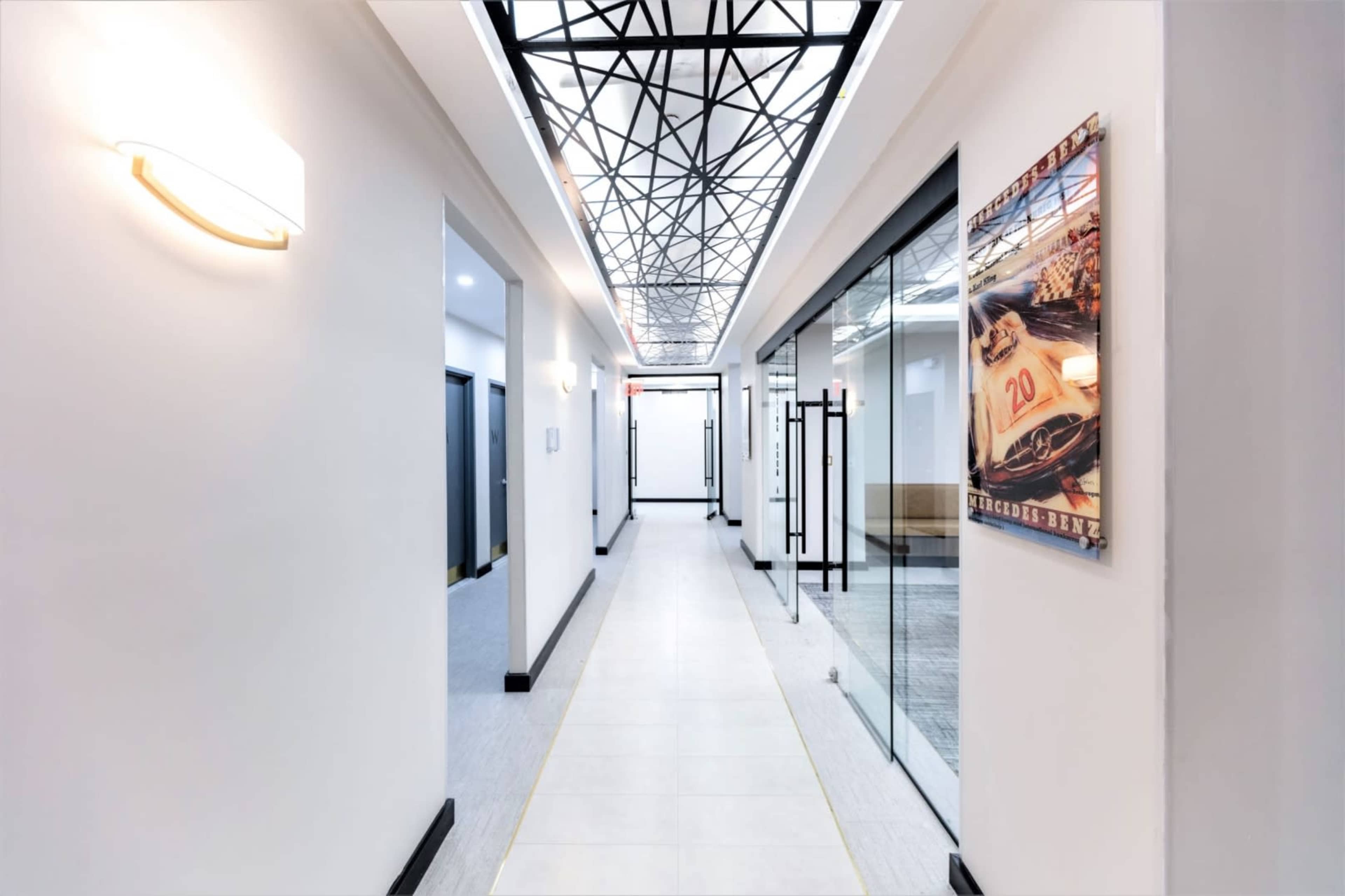 A well-lit corridor features glass doors on either side, leading to office spaces, with a geometric ceiling design and an automotive-themed artwork on one wall.