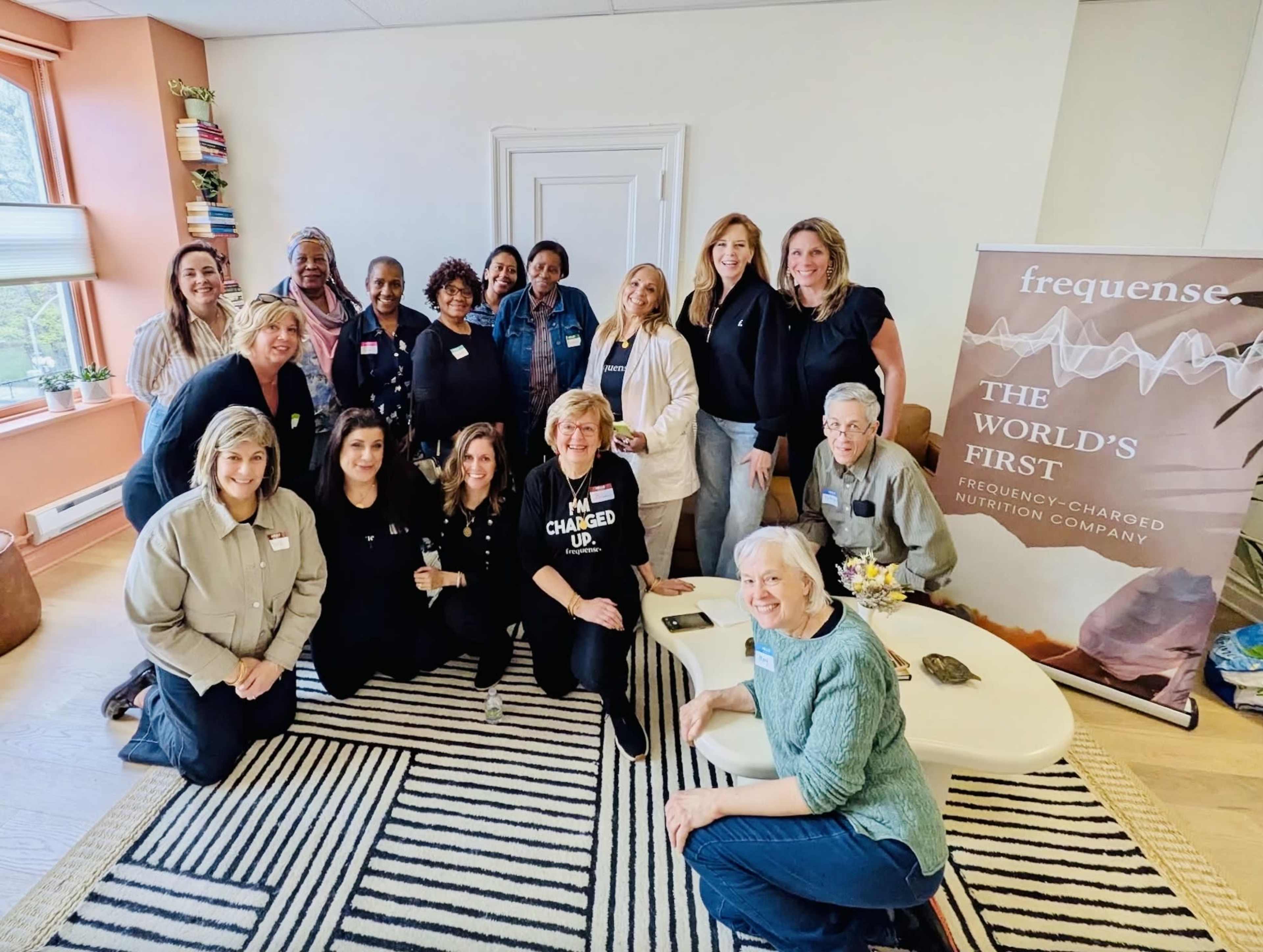A group of sixteen people poses together in a bright room, standing and sitting around a coffee table, with a banner that reads "frequences - THE WORLD'S FIRST" in the background.