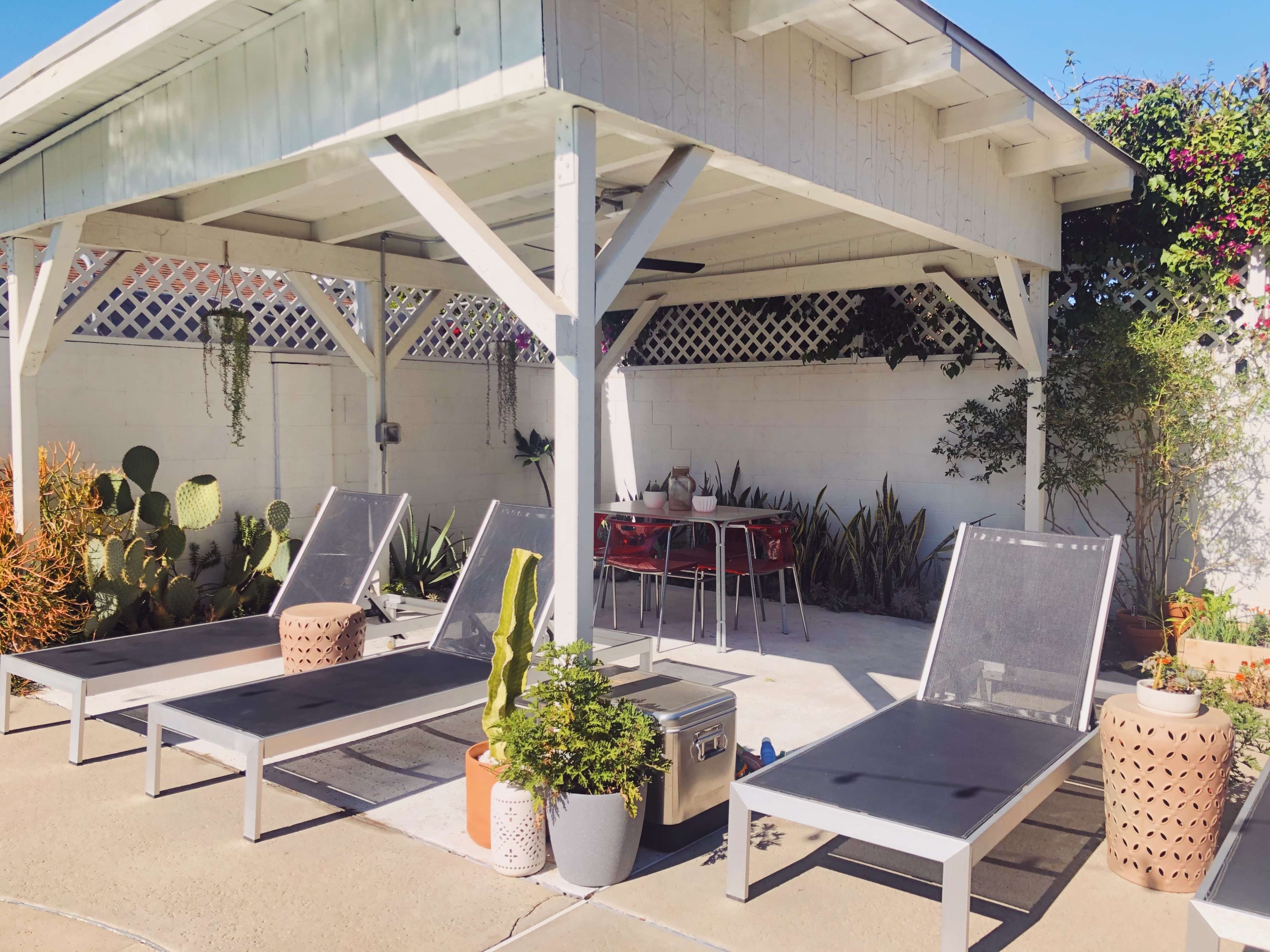 A shaded outdoor area with two lounge chairs, a small table set for four, and potted plants surrounded by a white wall.