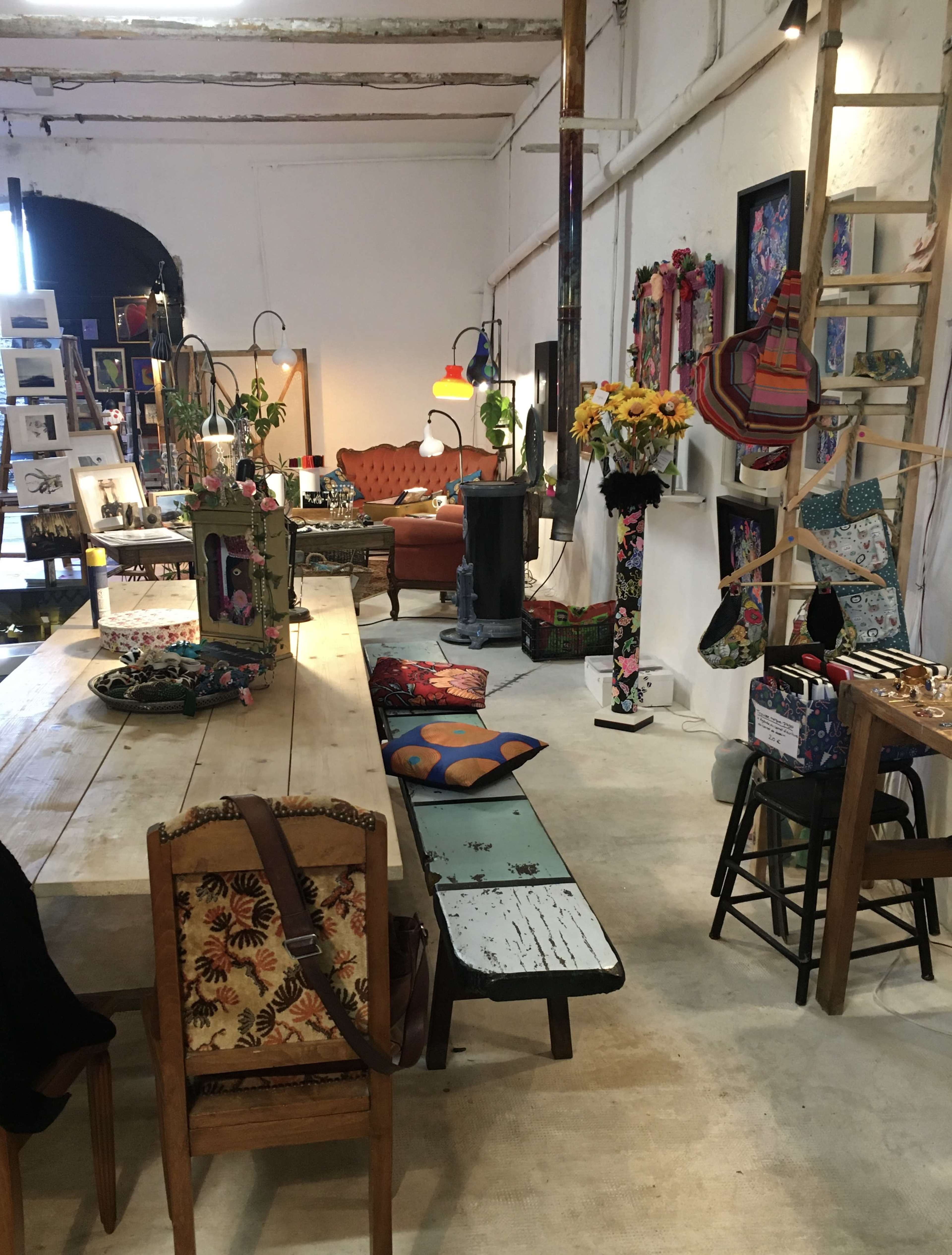 The image shows an eclectic interior of a creative space with various artworks, handcrafted items, and colorful decor arranged around a wooden table and seating.
