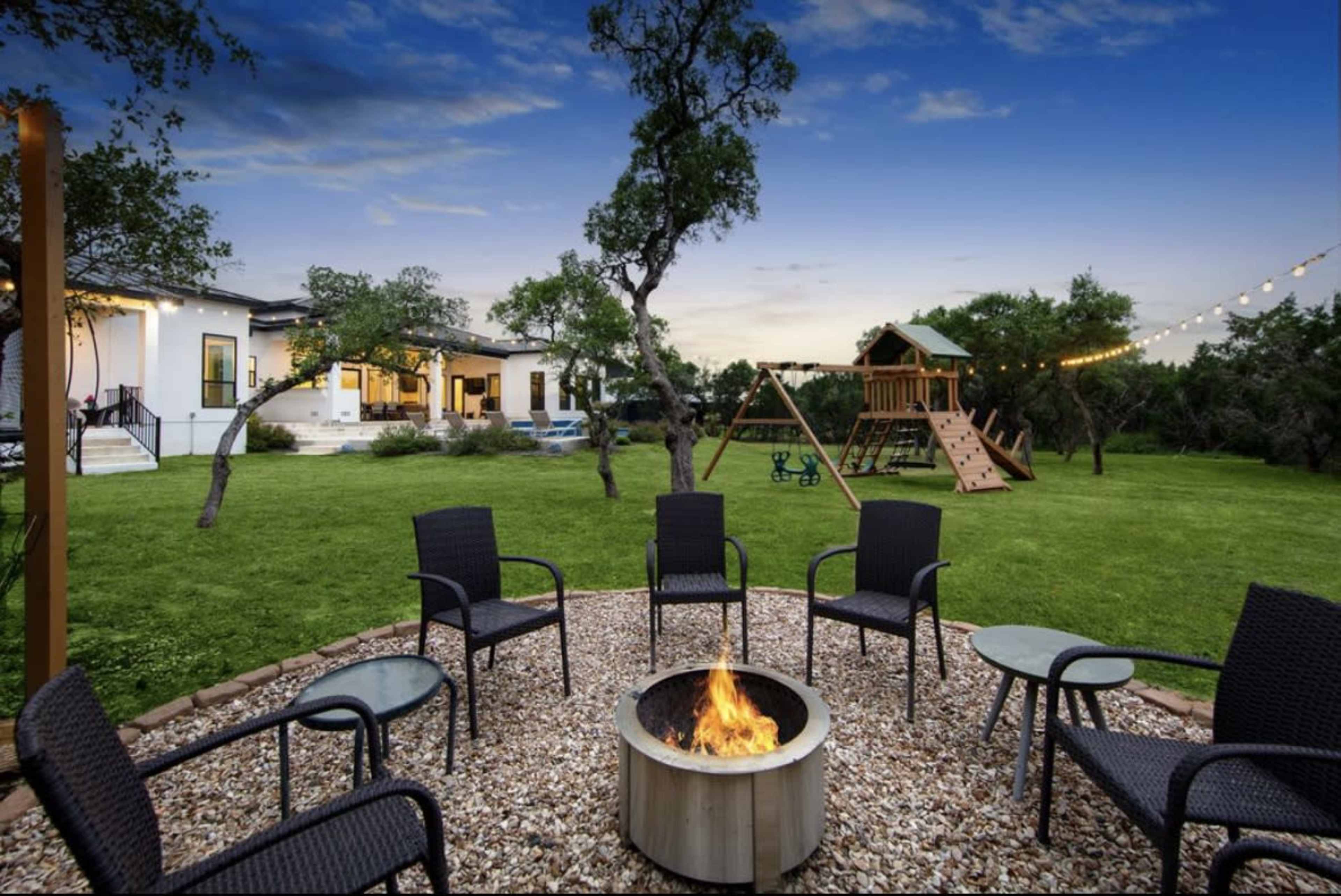 The image shows a backyard scene with a fire pit surrounded by chairs, a playground in the background, and a house partially visible with a pool.