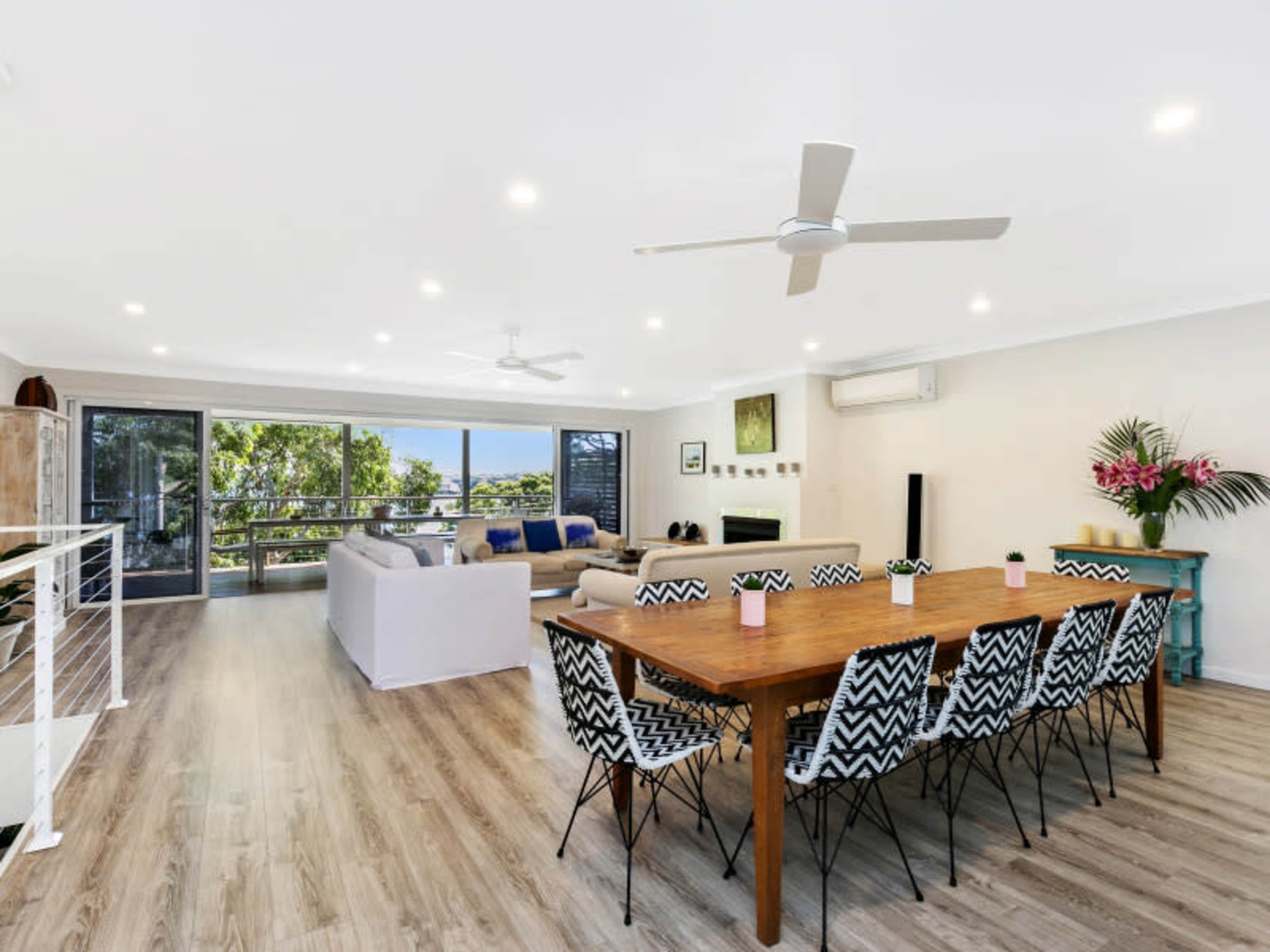 A spacious, modern open-plan living area featuring a dining table with patterned chairs, a seating area with a light-colored sofa, and large windows overlooking greenery.