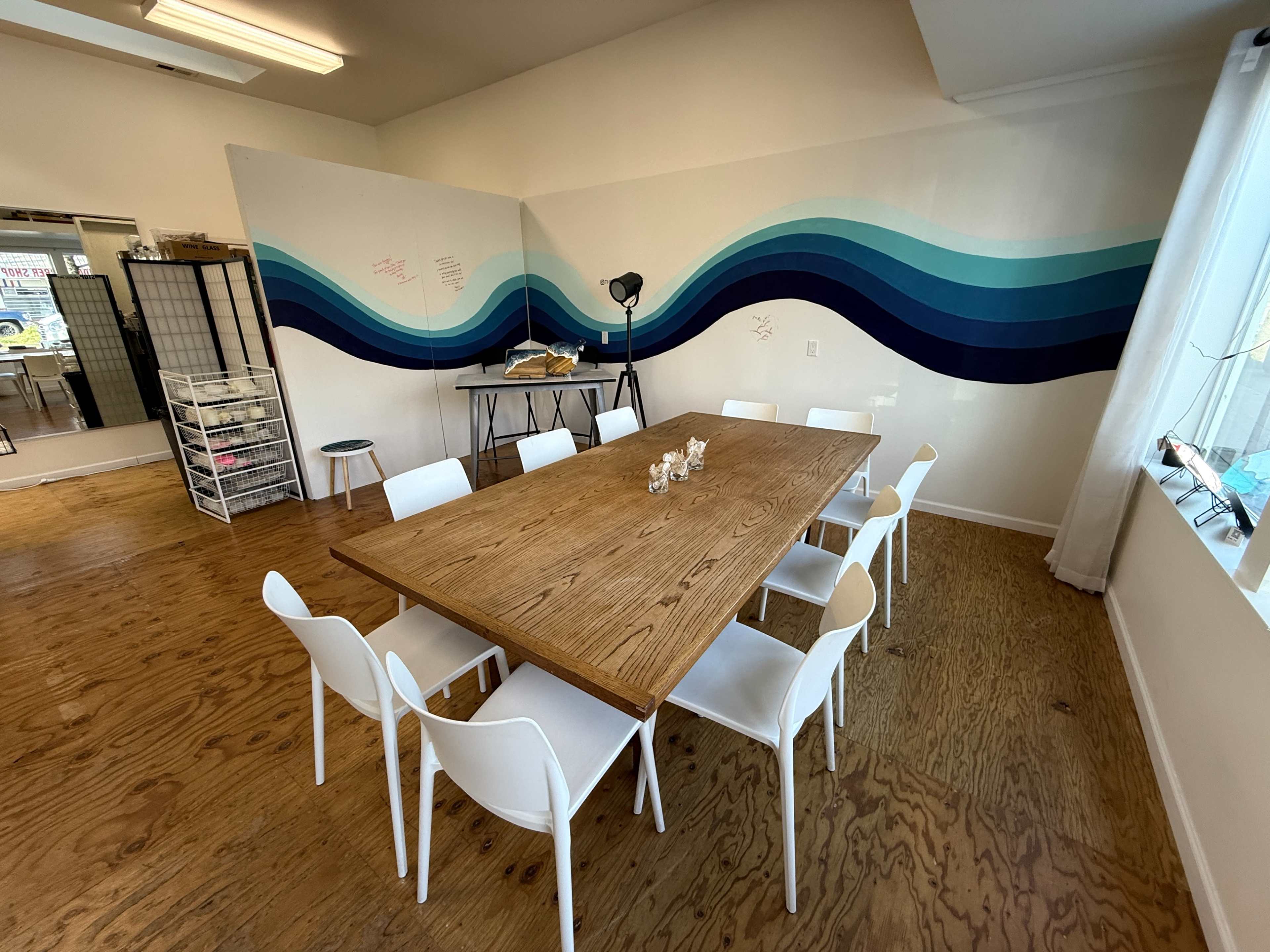 A large wooden table is surrounded by white chairs in a room featuring a blue wave mural on the wall.