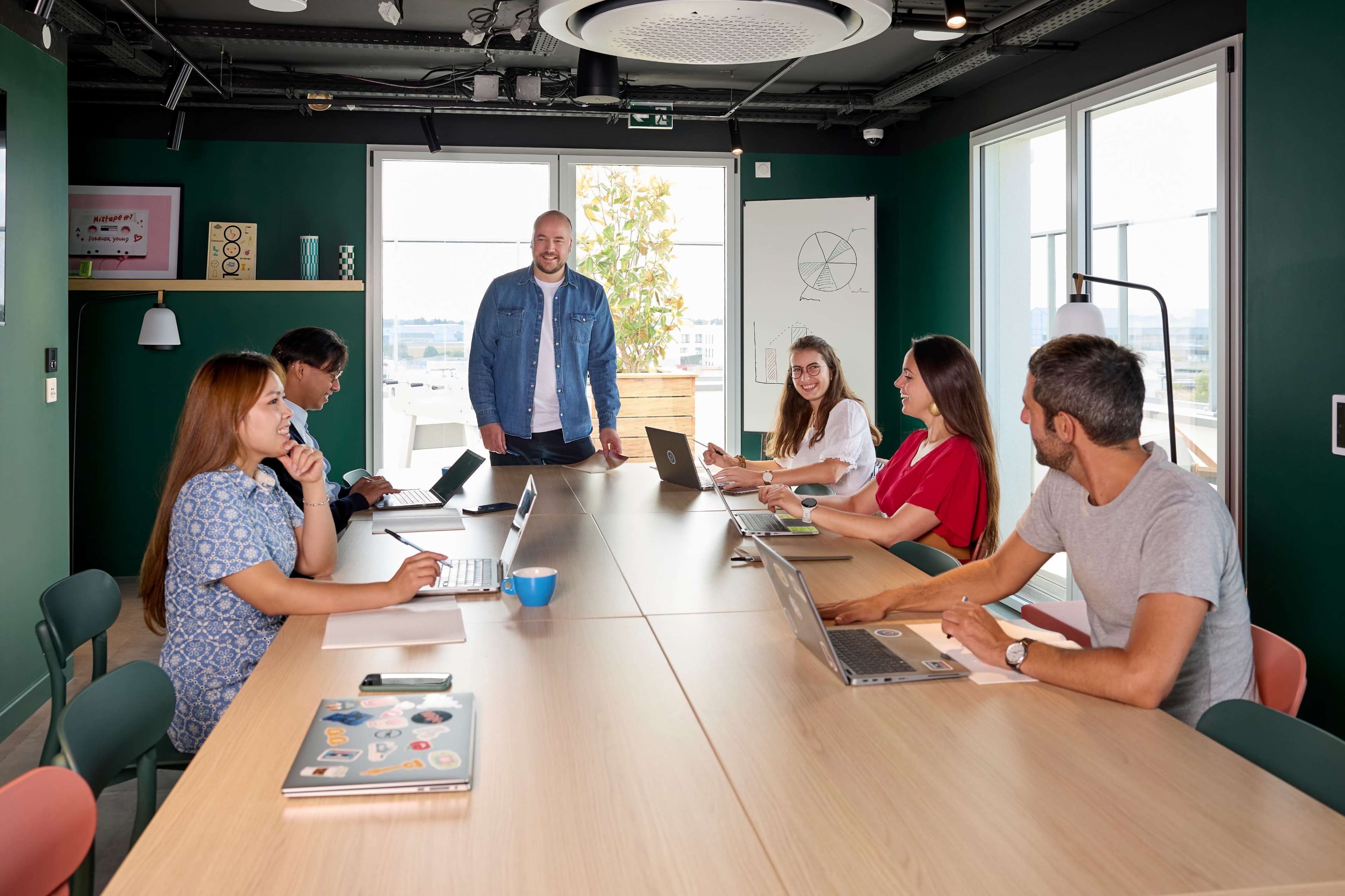 A group of six individuals engage in a discussion around a long table in a modern conference room with large windows and green walls.