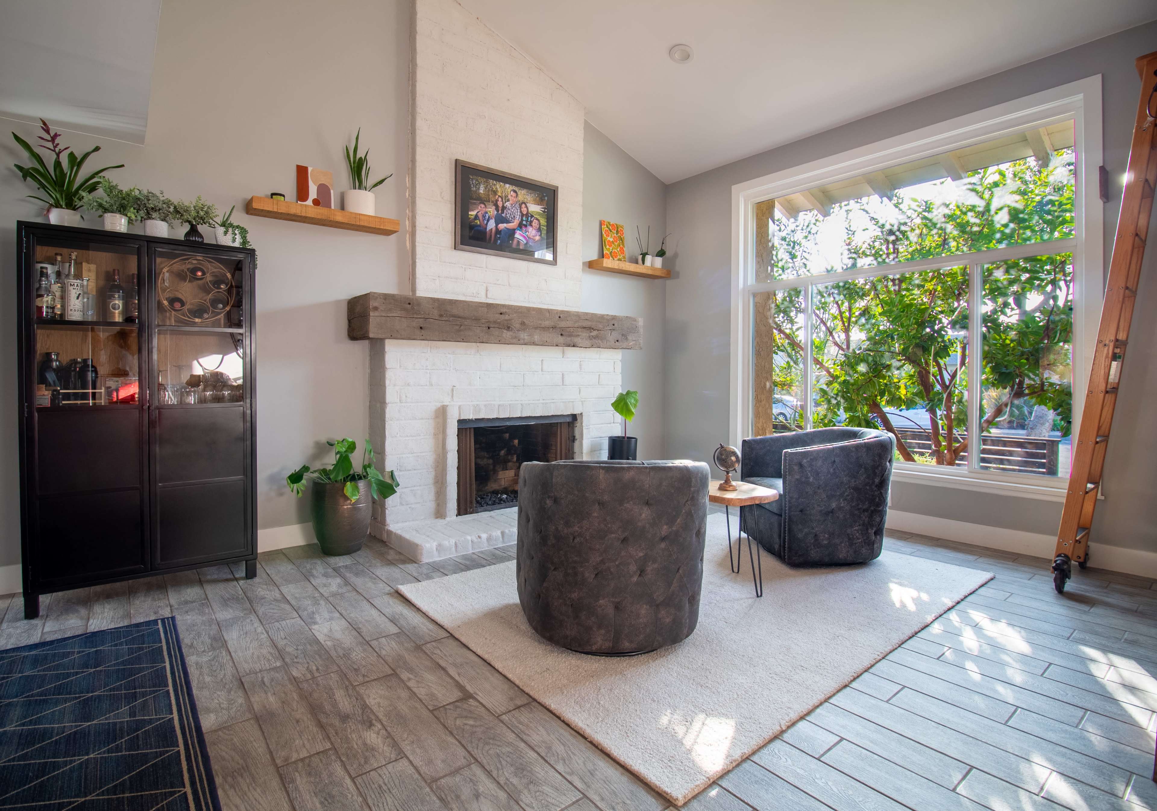 A modern living room featuring a fireplace with a wooden mantel, two chairs, a small side table, and a large window that allows natural light to illuminate the space.