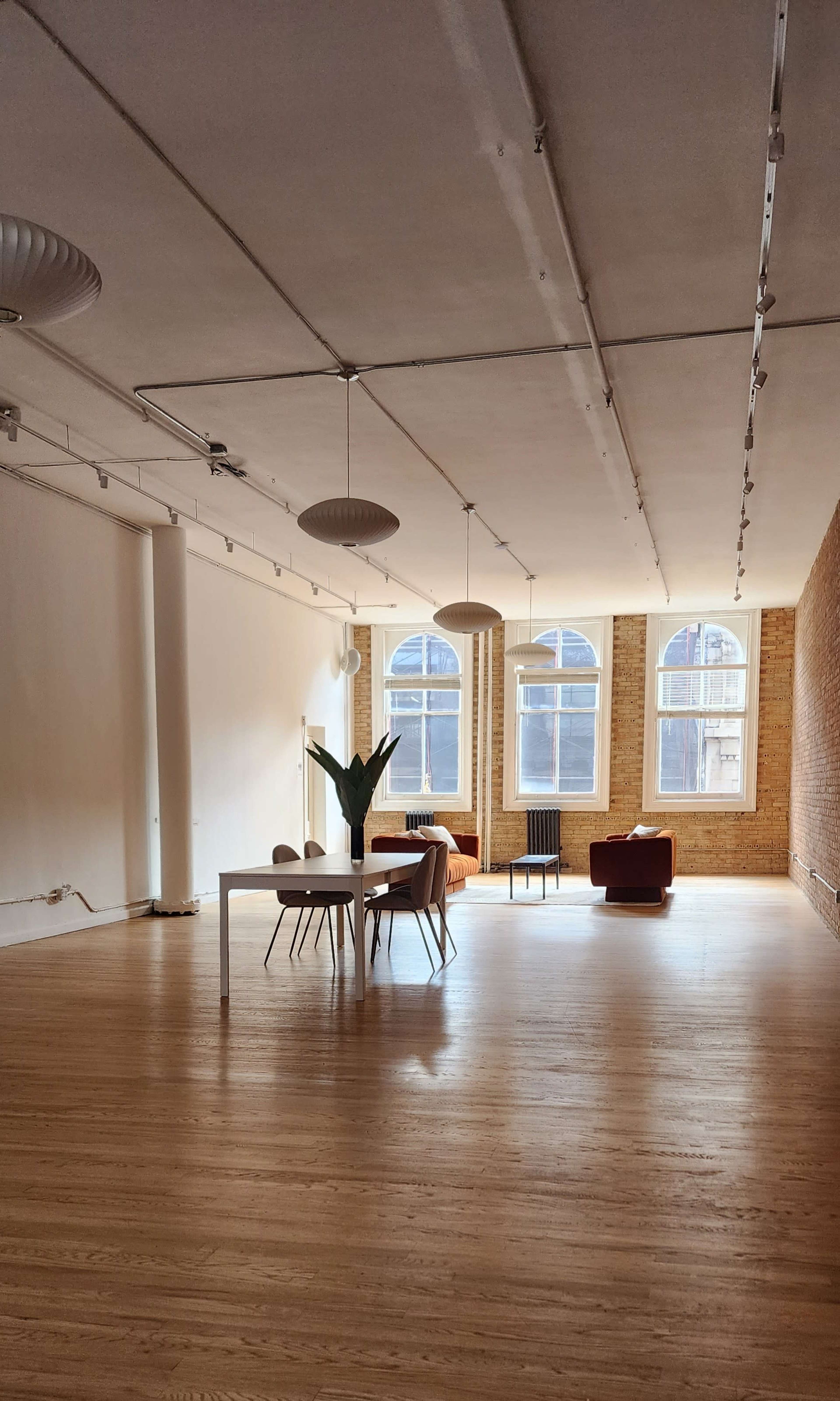 The image shows a spacious, well-lit room with wooden flooring, featuring a table with chairs and a small seating area beside a brick wall.