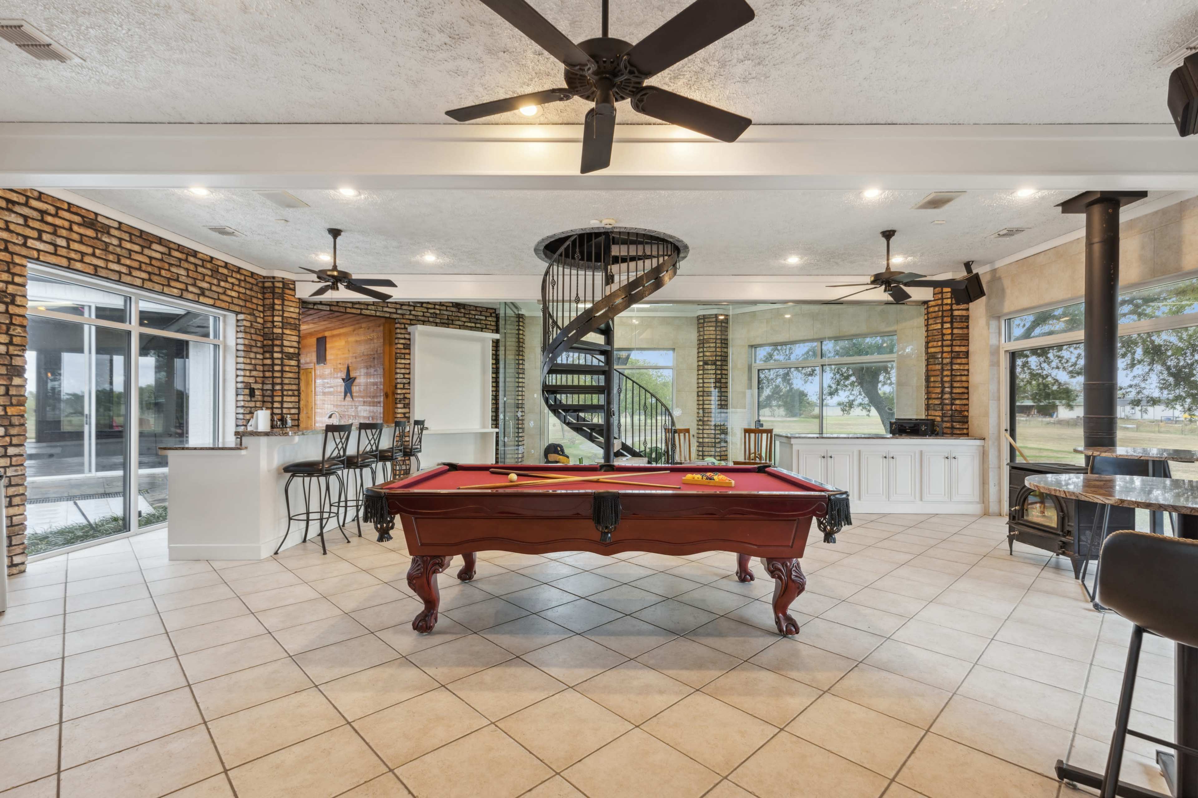 The image shows a spacious indoor lounge area with a pool table in the center, surrounded by large windows and a spiral staircase, featuring a bar counter and a brick accent wall.