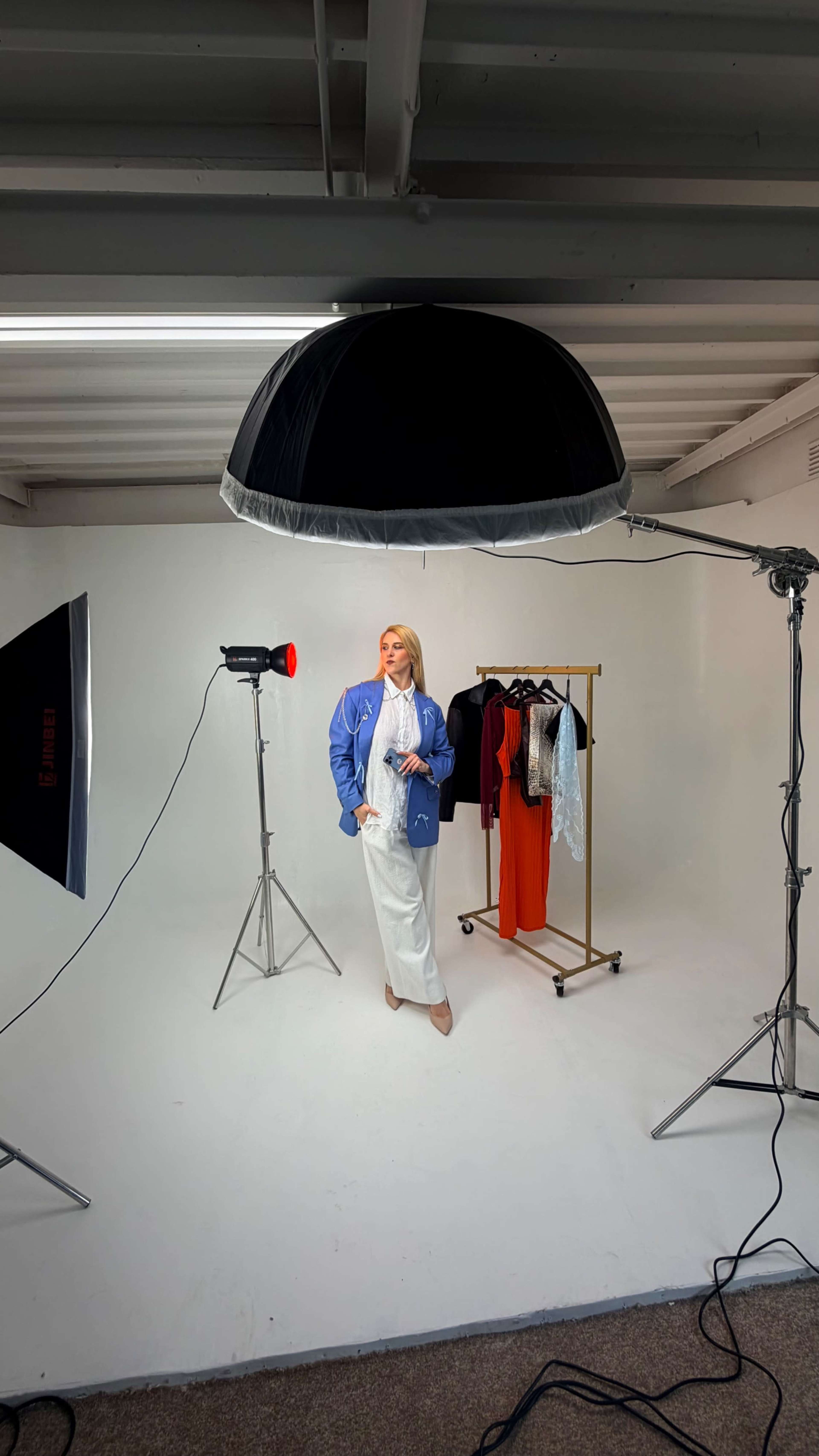 A model poses in a studio setting, standing in front of a clothing rack with a variety of garments while under bright studio lights.