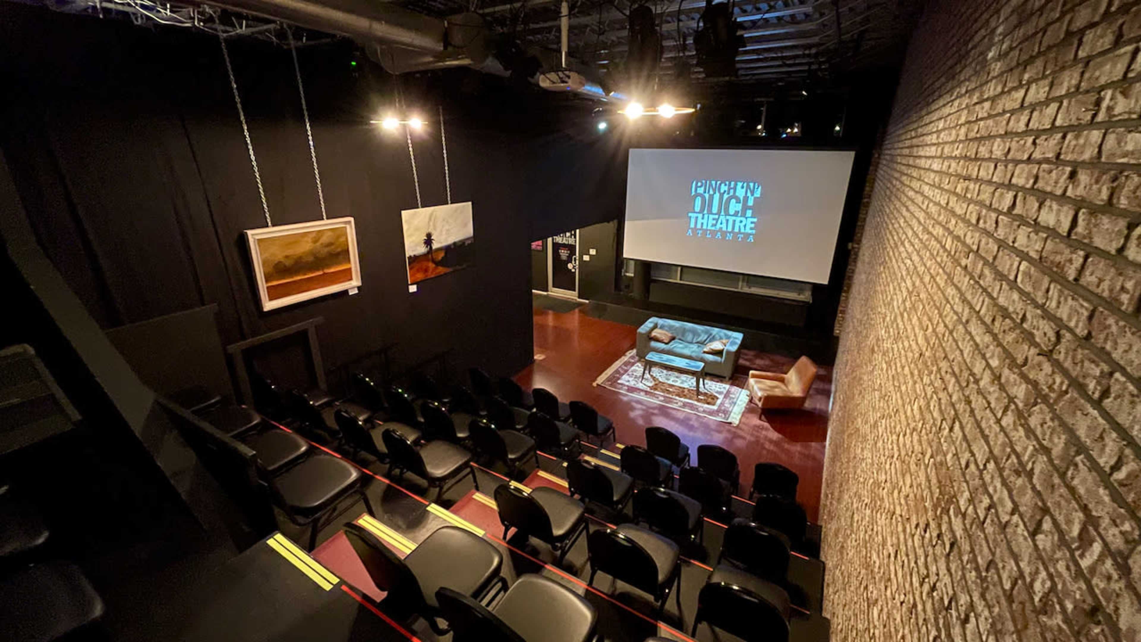 The image shows a small theater space with black seating and a large screen at the front, surrounded by brick walls and dim lighting.