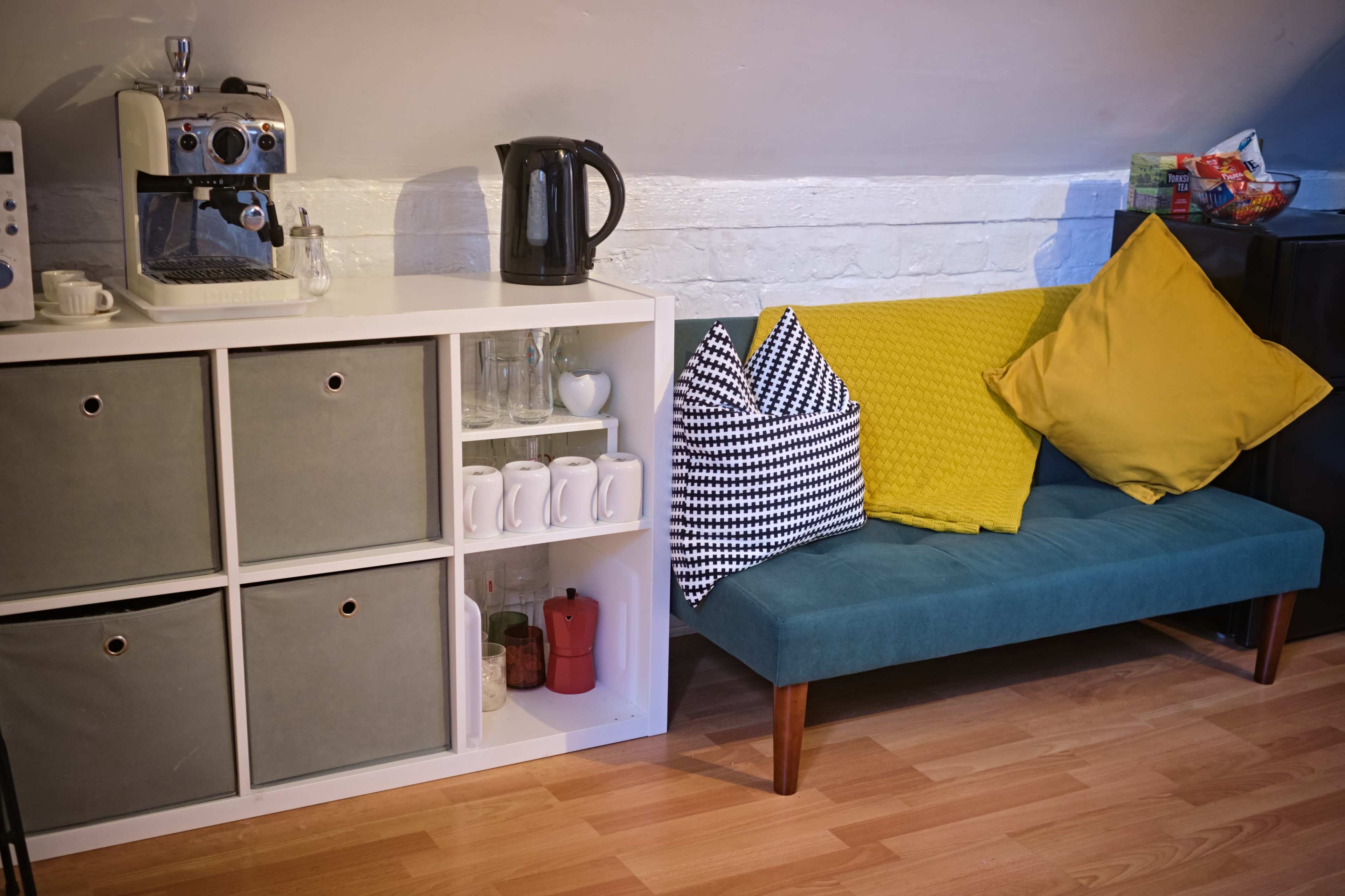A teal couch with colorful pillows is positioned next to a white shelving unit containing glassware and storage bins, alongside a coffee maker and kettle.