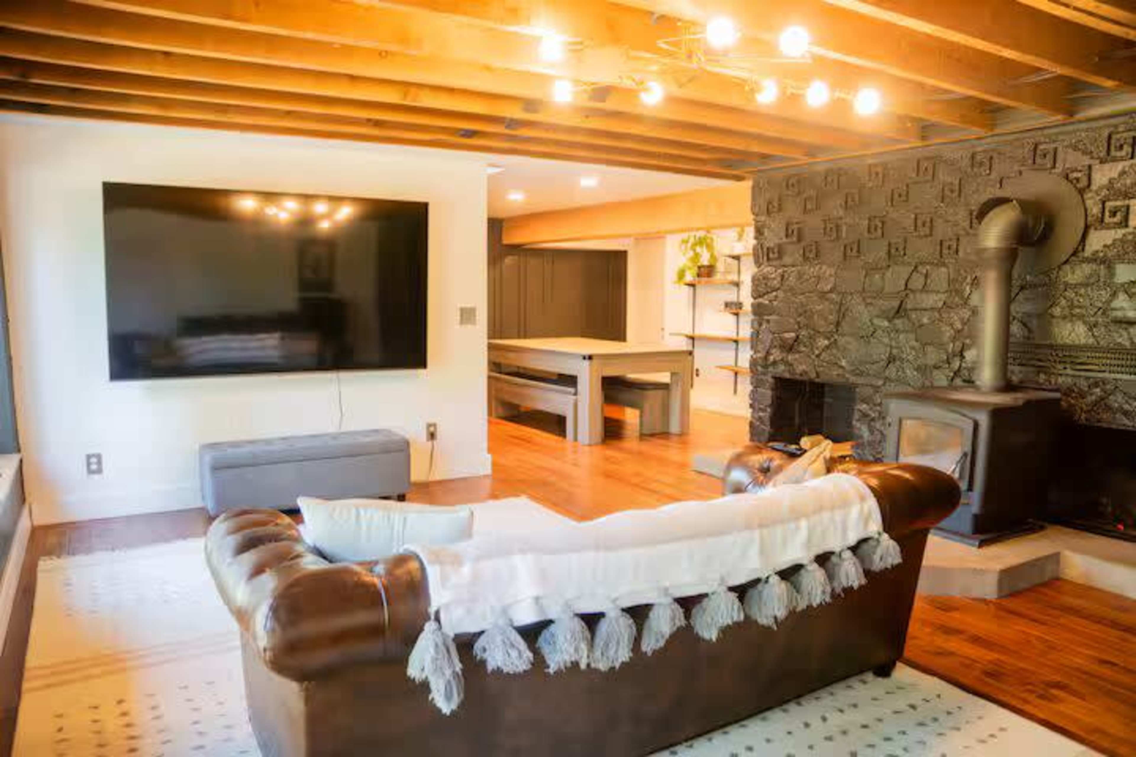 The image shows a modern living room with a large wall-mounted television, a leather sofa with a throw blanket, and a stone fireplace.