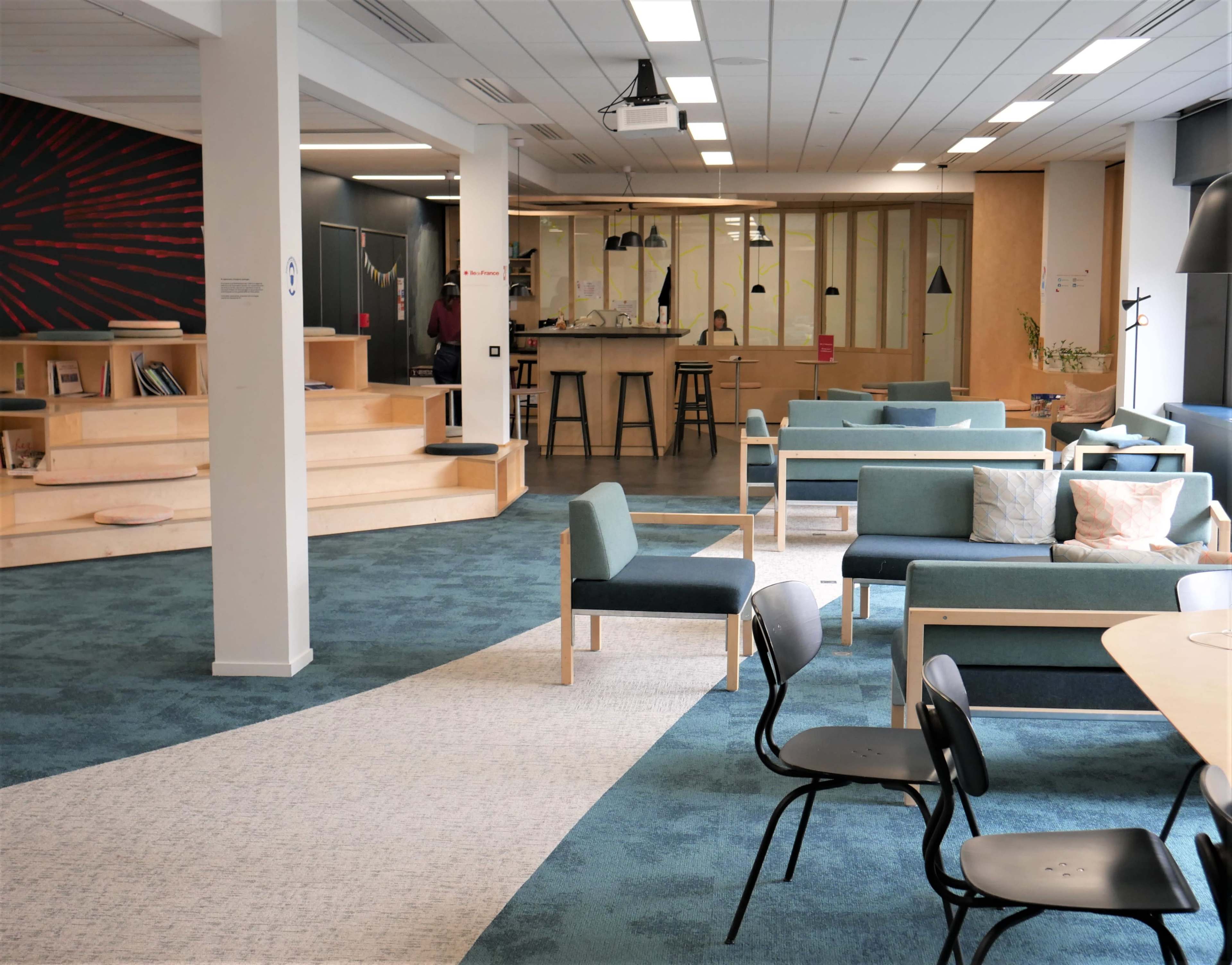 The image shows a modern, open common area with seating arrangements, a light blue carpet, and a counter area in the background.