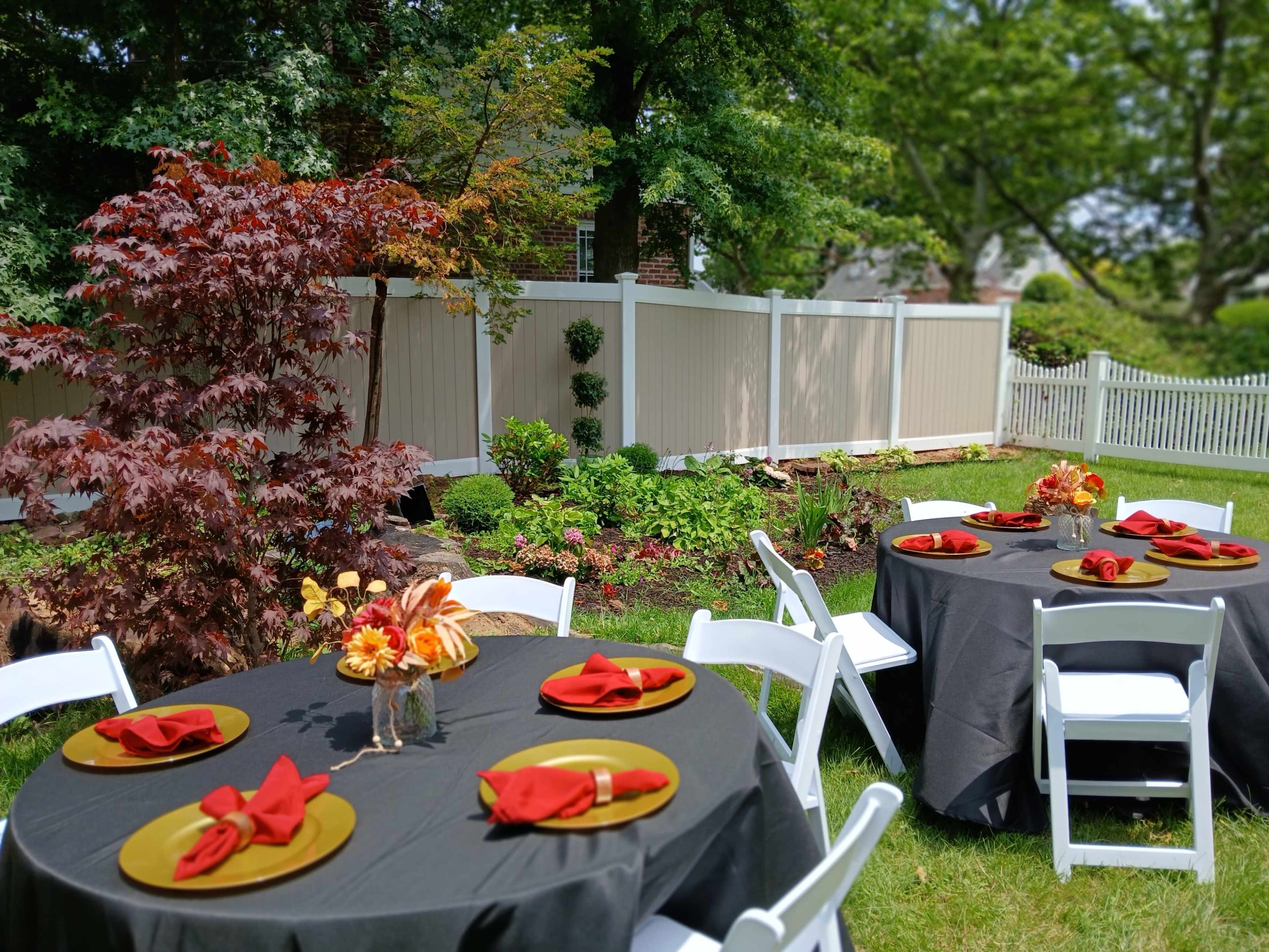 A backyard setting features round tables with black tablecloths, red napkins, and floral centerpieces, surrounded by greenery and a white picket fence.