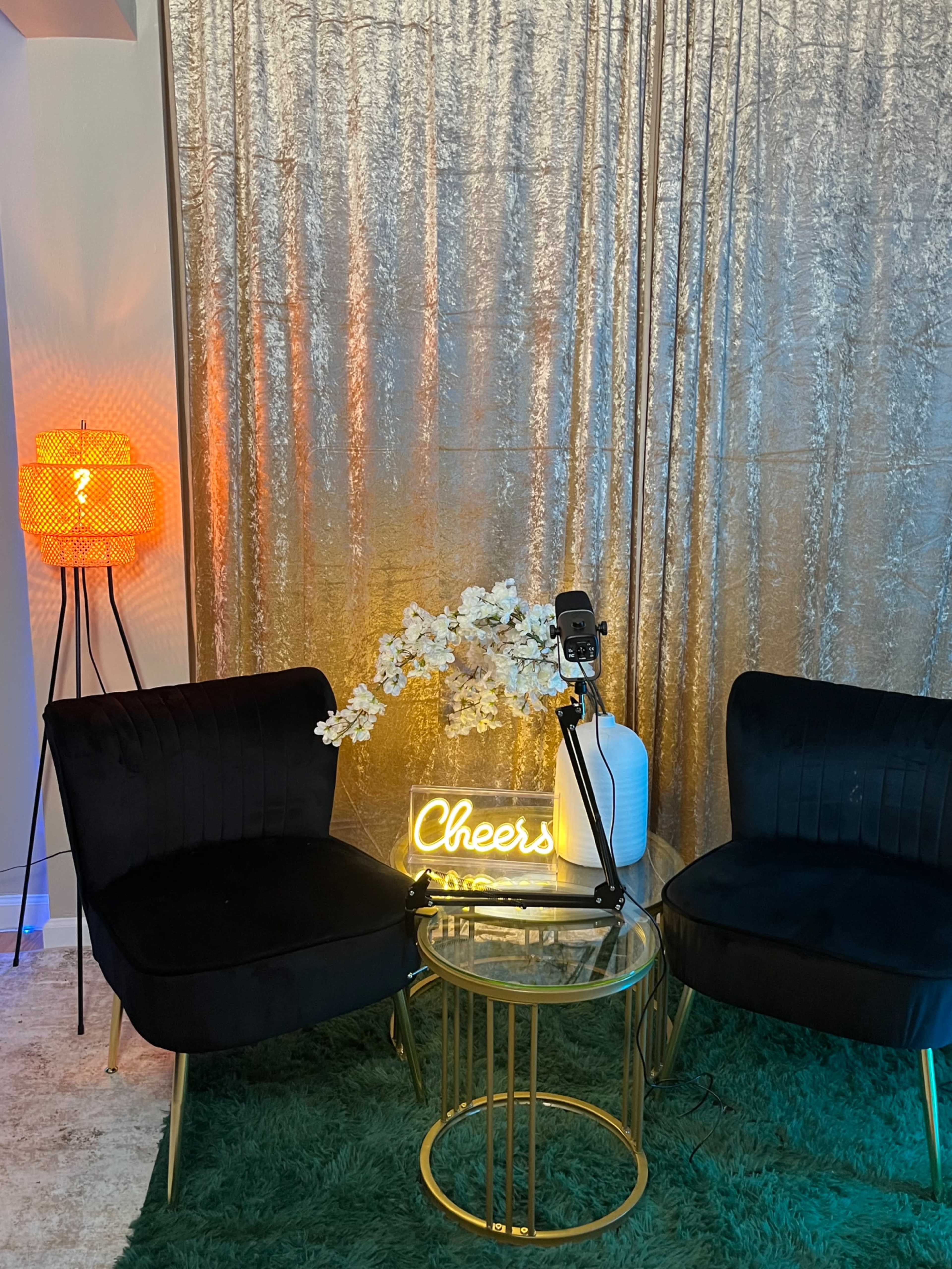 The scene features two black velvet chairs surrounding a glass table, with a neon "Cheers" sign and a flower arrangement in front of a shimmering curtain, alongside a decorative lamp.