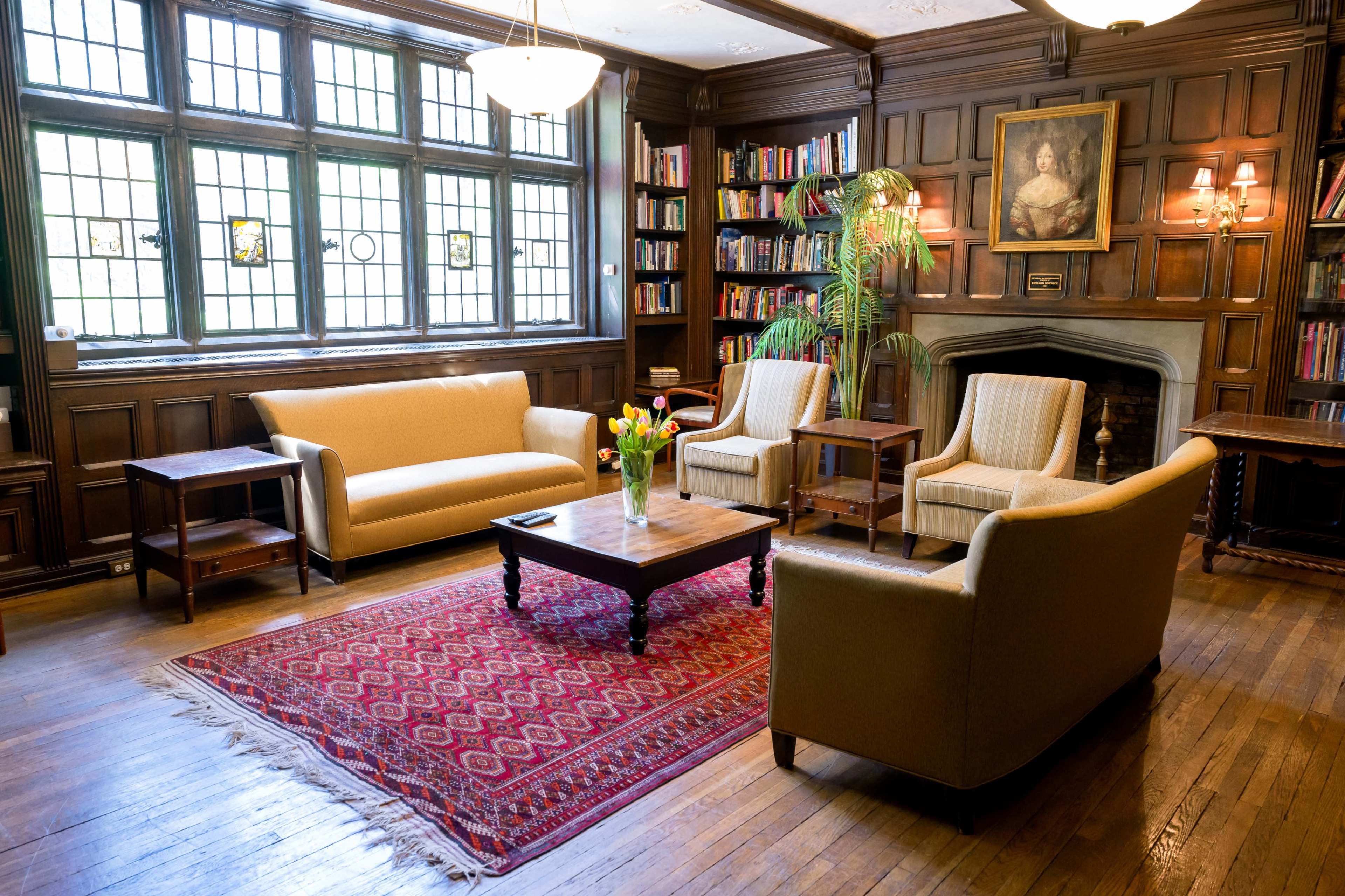A cozy living room with wooden paneling, featuring a sofa, two armchairs, a coffee table, and a colorful area rug, alongside a bookshelf and a fireplace.