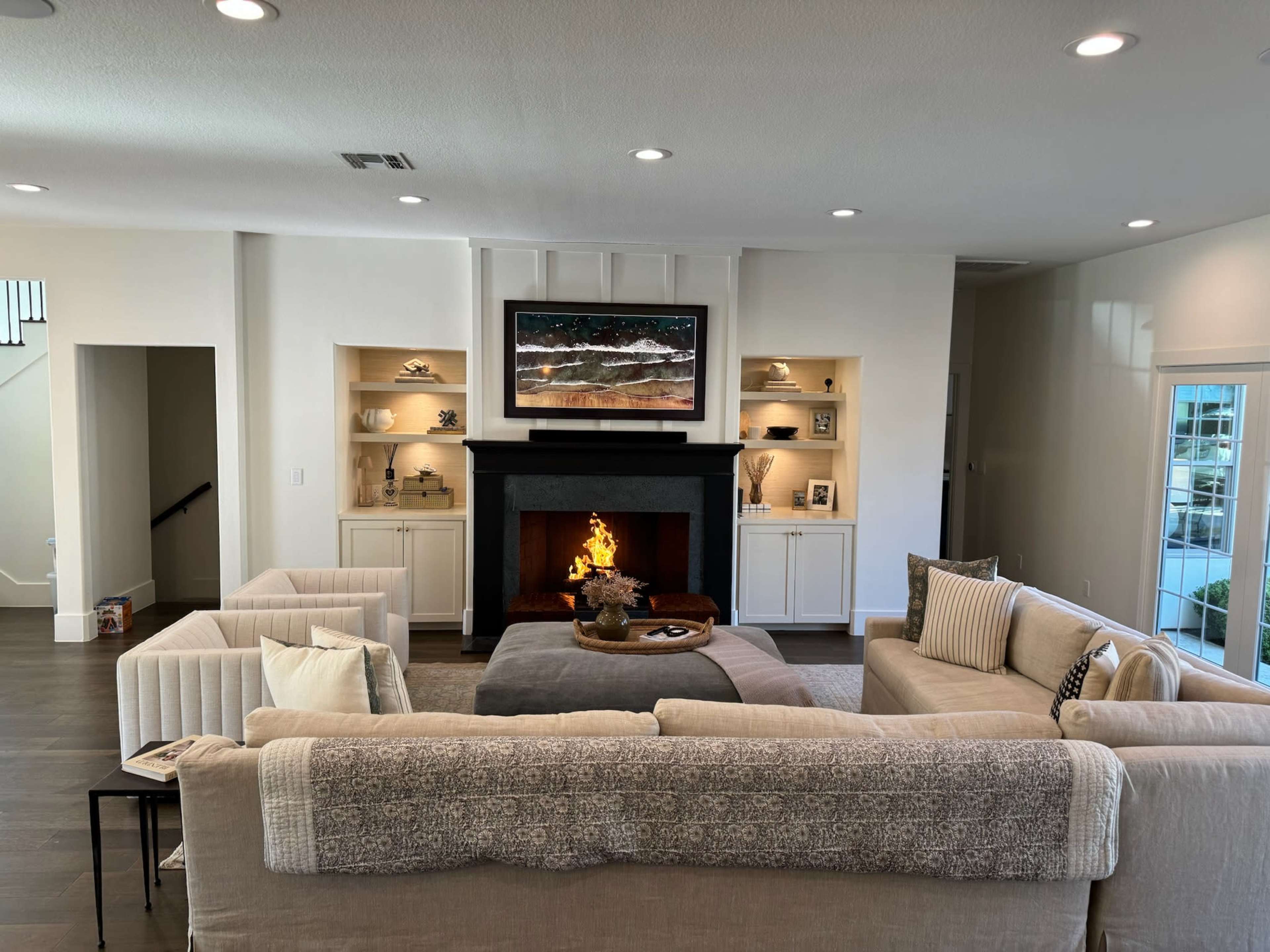 A modern living room with a fireplace, a large sofa, and two armchairs arranged around a coffee table in front of a television mounted above the mantel.