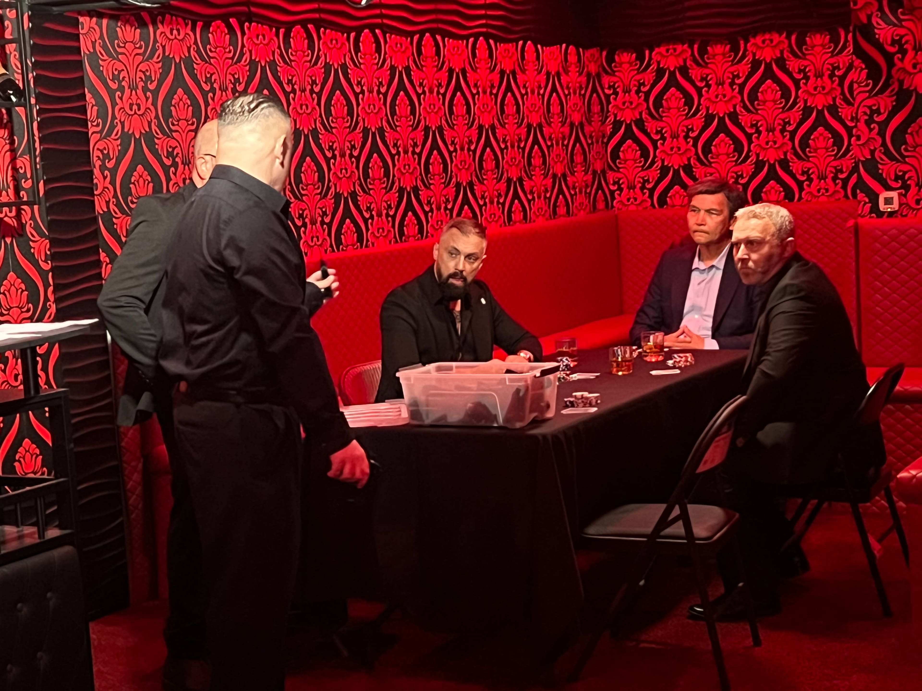 A group of four men is seated around a table in a dimly lit room with red wallpaper, while a man stands nearby, appearing to discuss something with them.