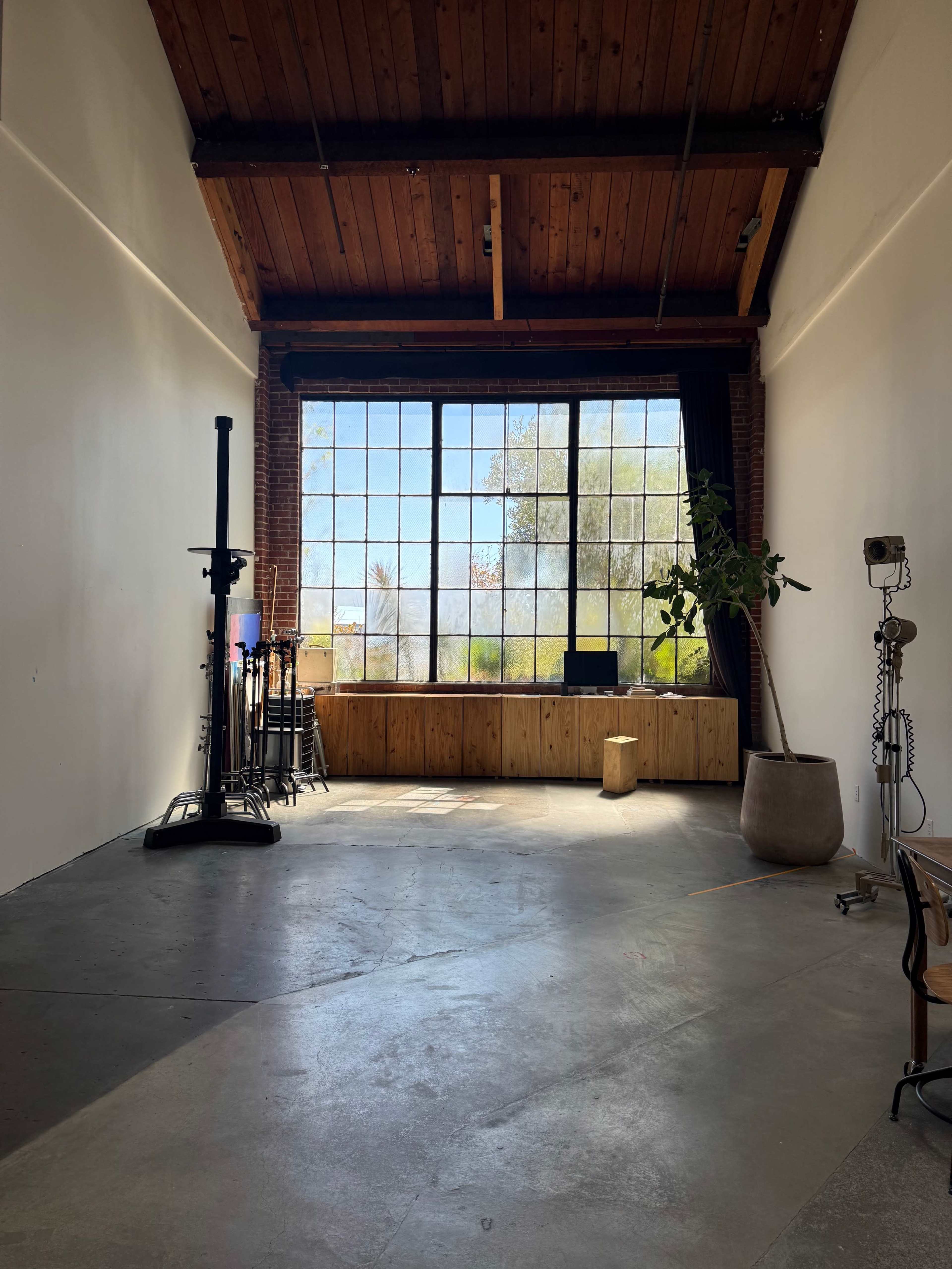 The image shows a spacious room with a large window, wooden ceiling beams, and a wooden counter, featuring a potted plant and various studio equipment.