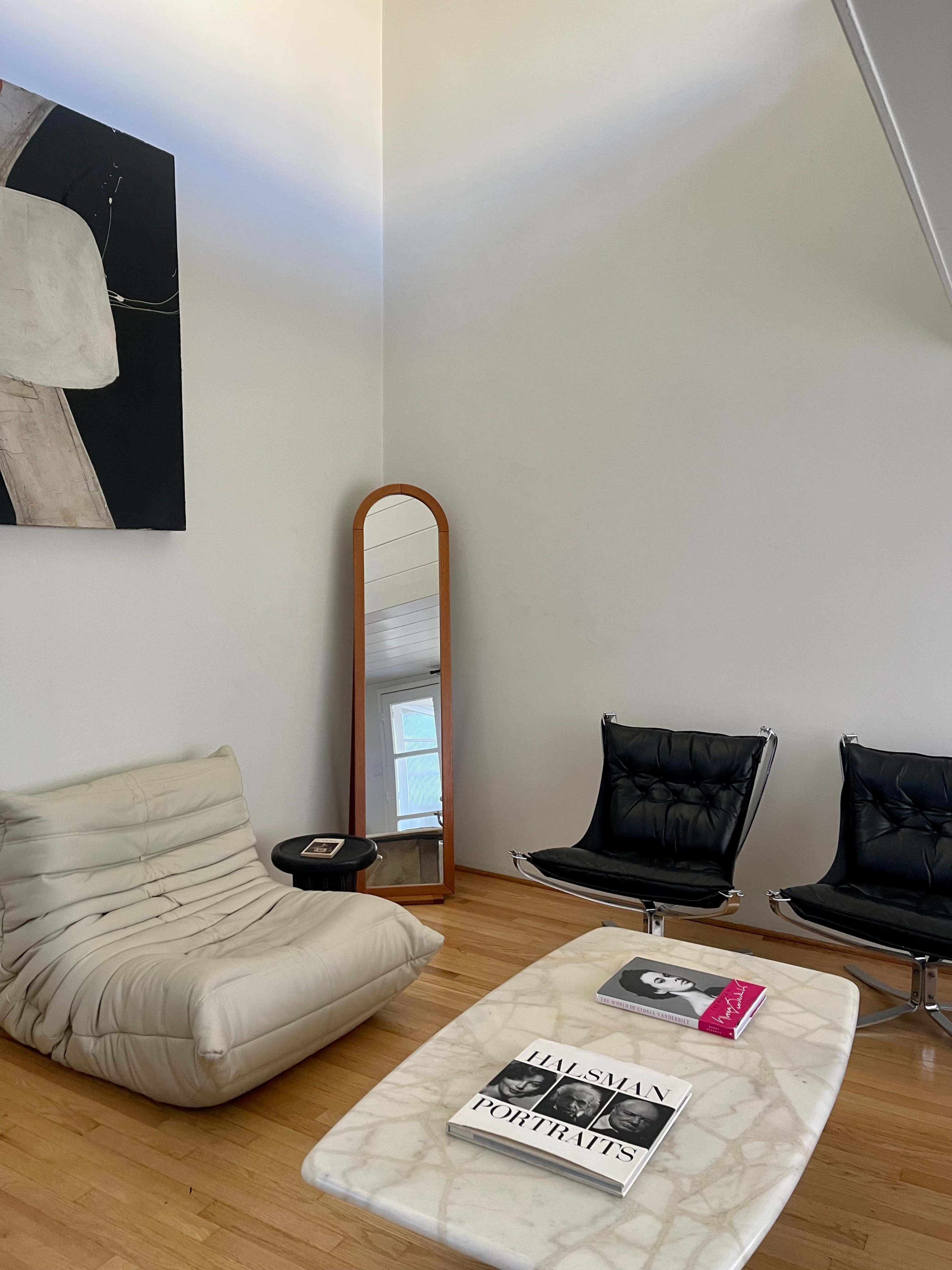 A minimalist living room with a white lounge chair, two black armchairs, a marble coffee table, and a tall mirror against a wall adorned with abstract art.