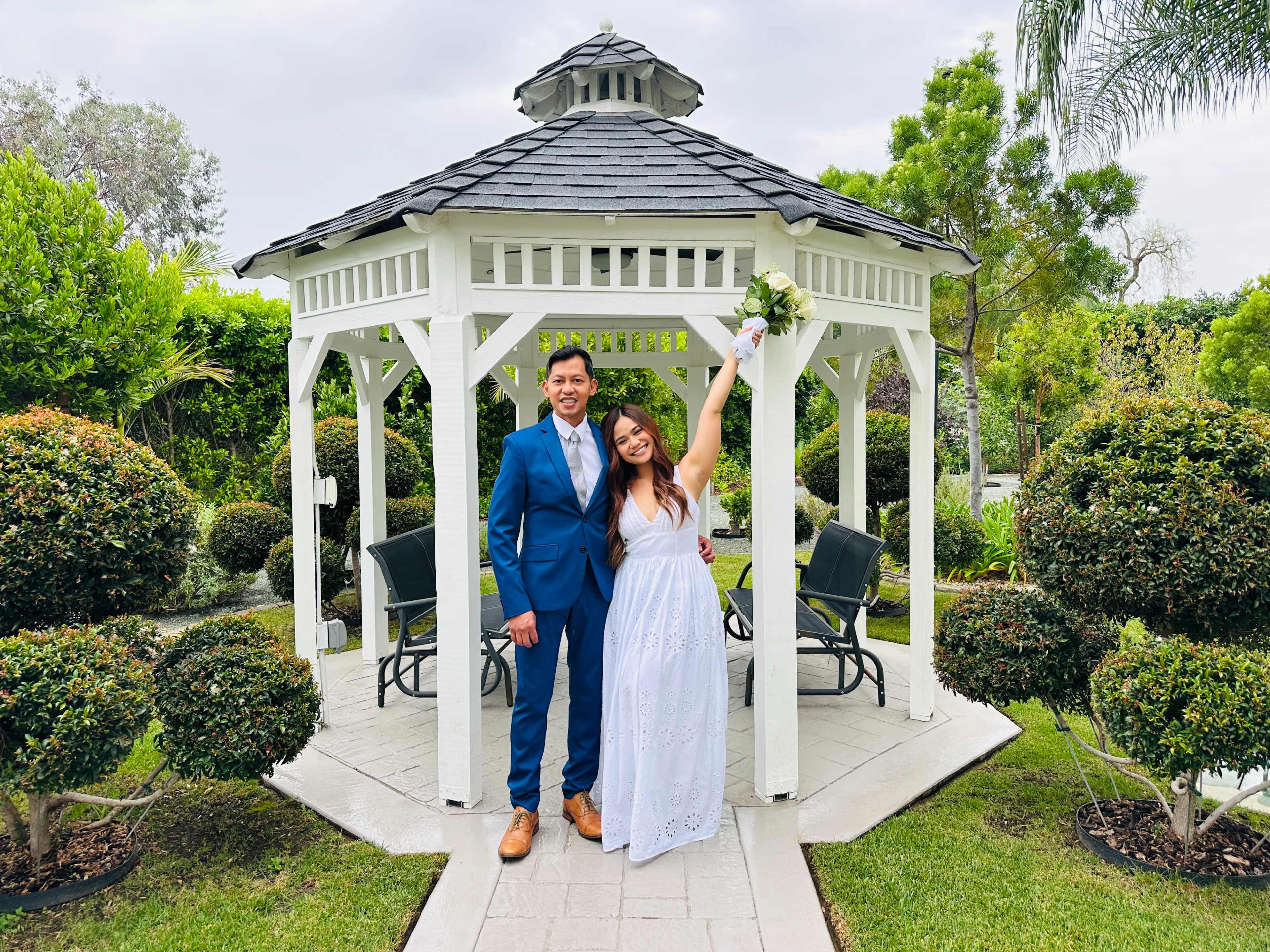 A couple stands together under a gazebo in a landscaped garden, with the woman holding a bouquet and wearing a white dress, while the man is dressed in a blue suit.