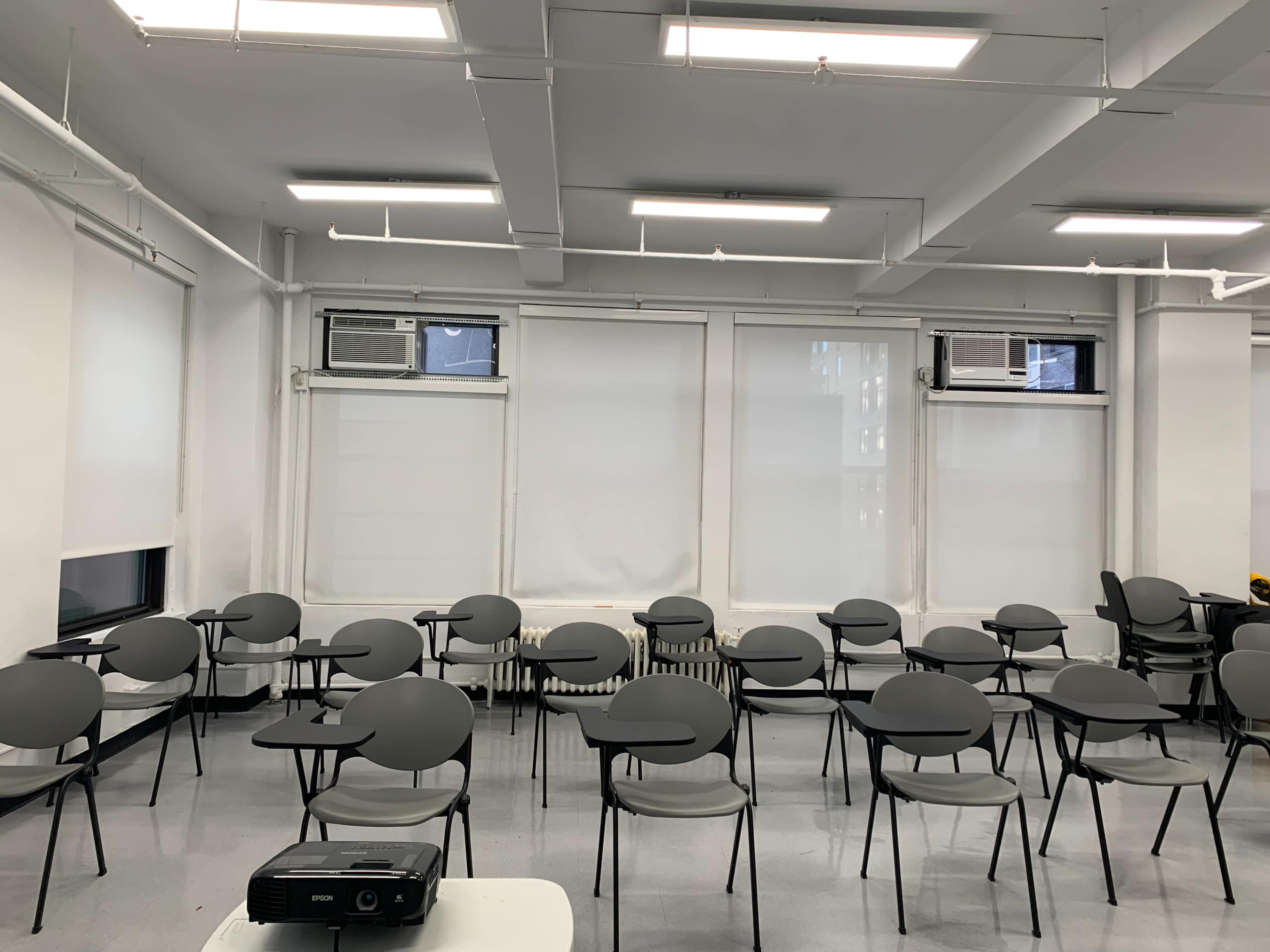 A classroom is arranged with rows of empty gray chairs and a projector on a table.
