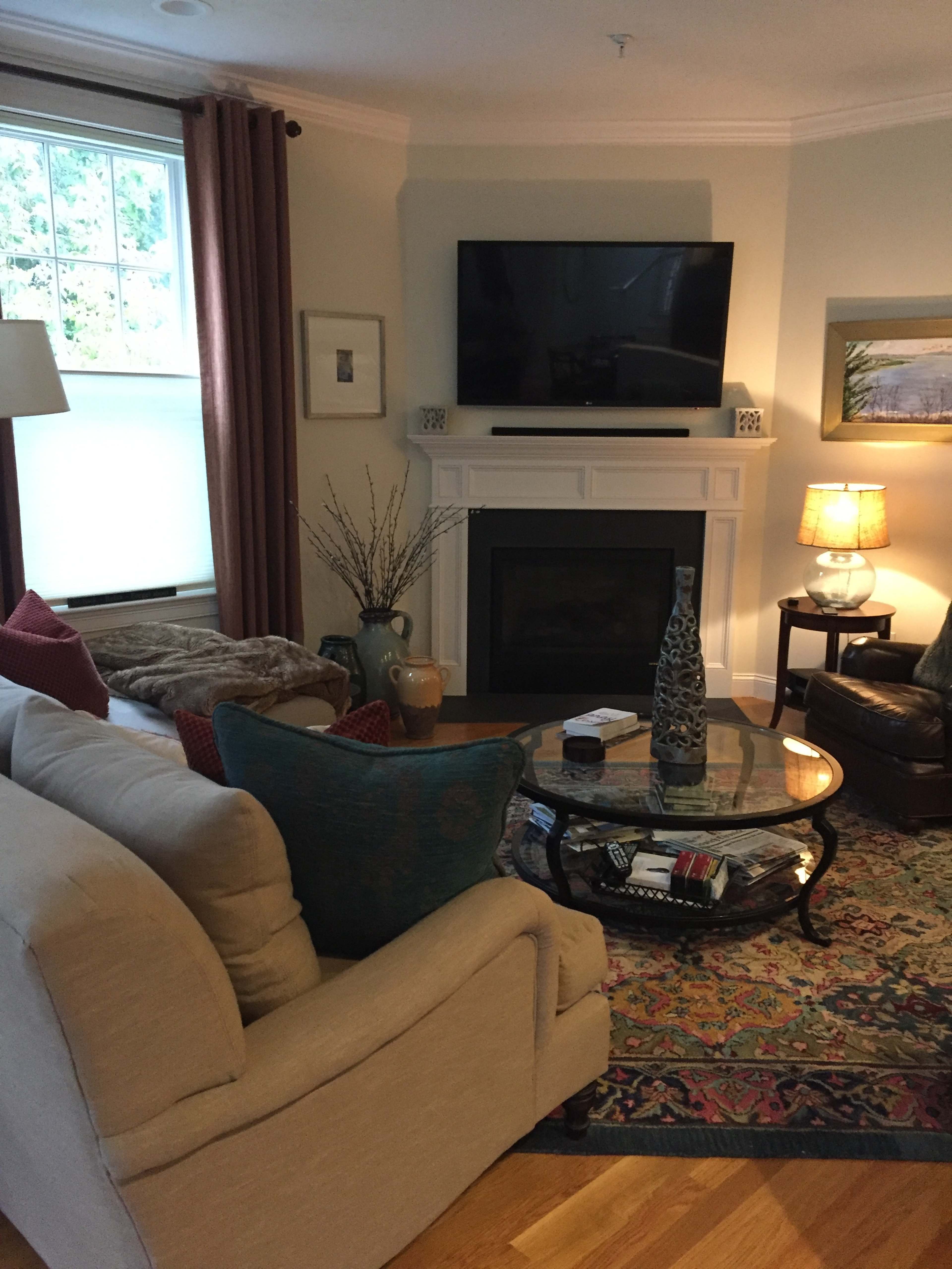 The living room features a beige sofa, an armchair, and a coffee table centered in front of a fireplace with a large flat-screen TV mounted above it.