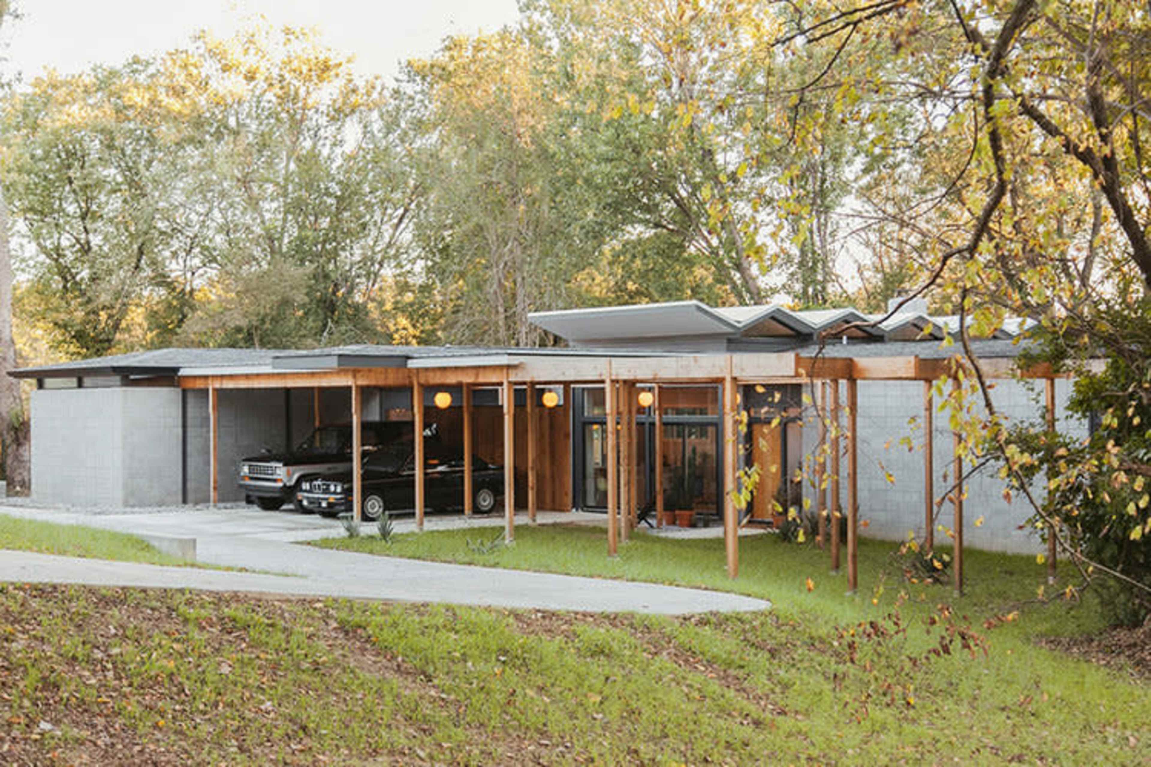 A modern single-story house features a flat roof, wooden columns, and a driveway leading to two parked cars.