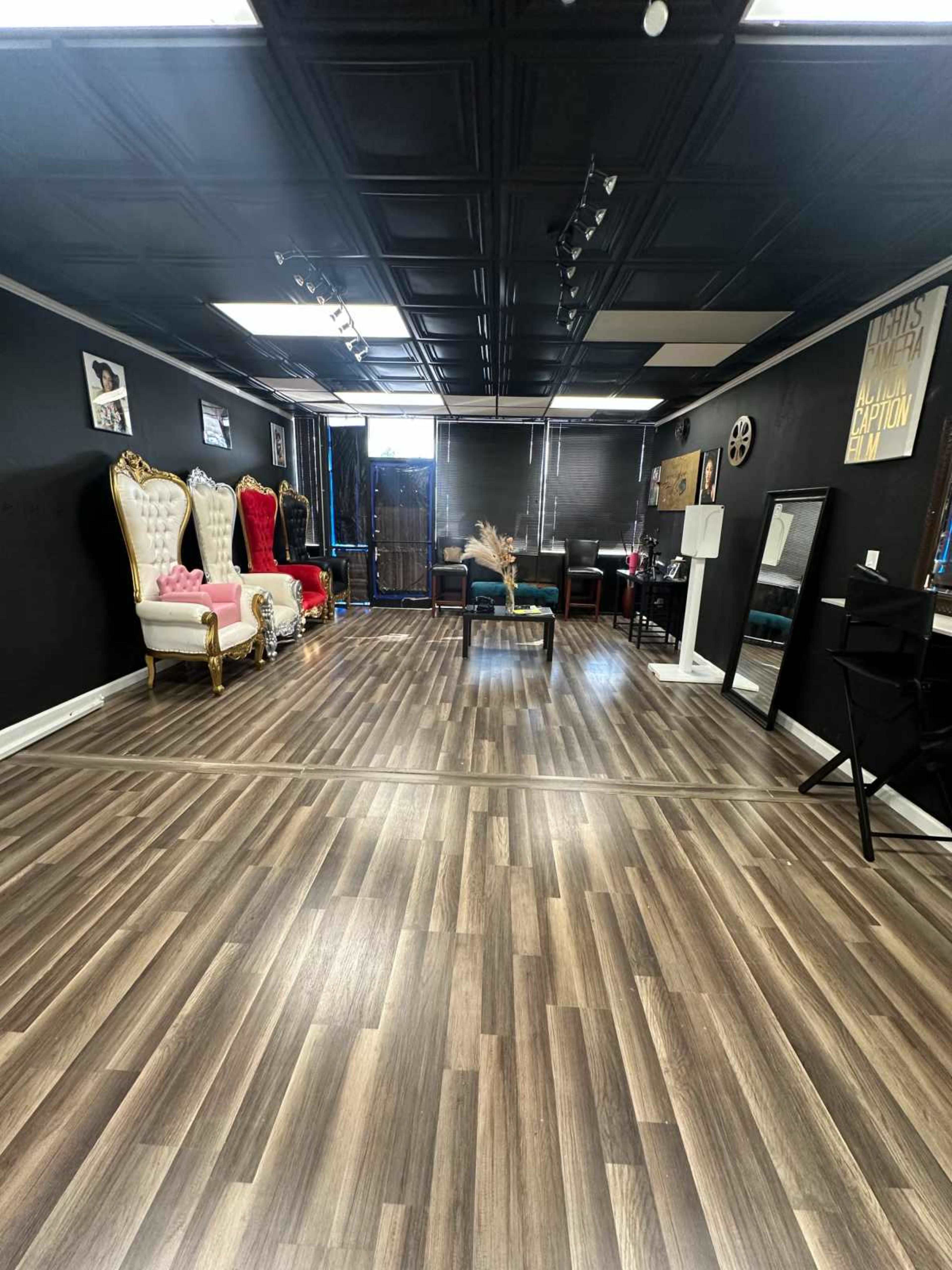 The image shows a spacious room with dark walls, wooden flooring, several ornate chairs, and natural light coming from the windows.