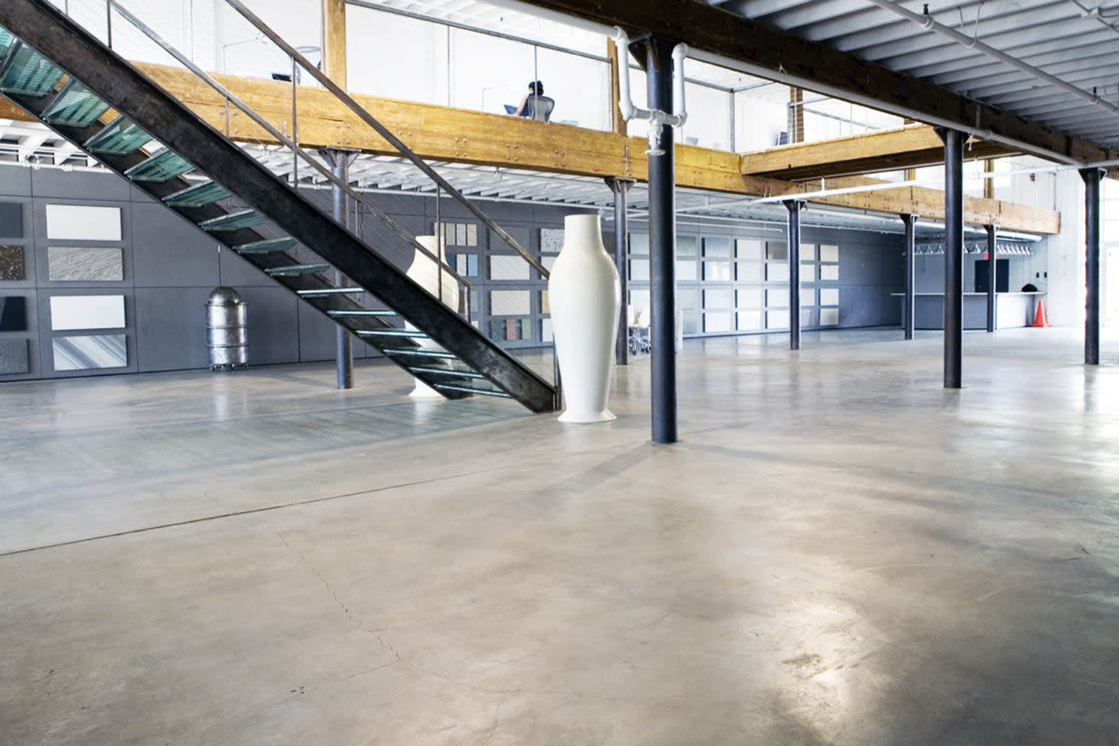 The image shows a spacious industrial interior featuring a staircase made of metal and wood, a large white vase, and a polished concrete floor.