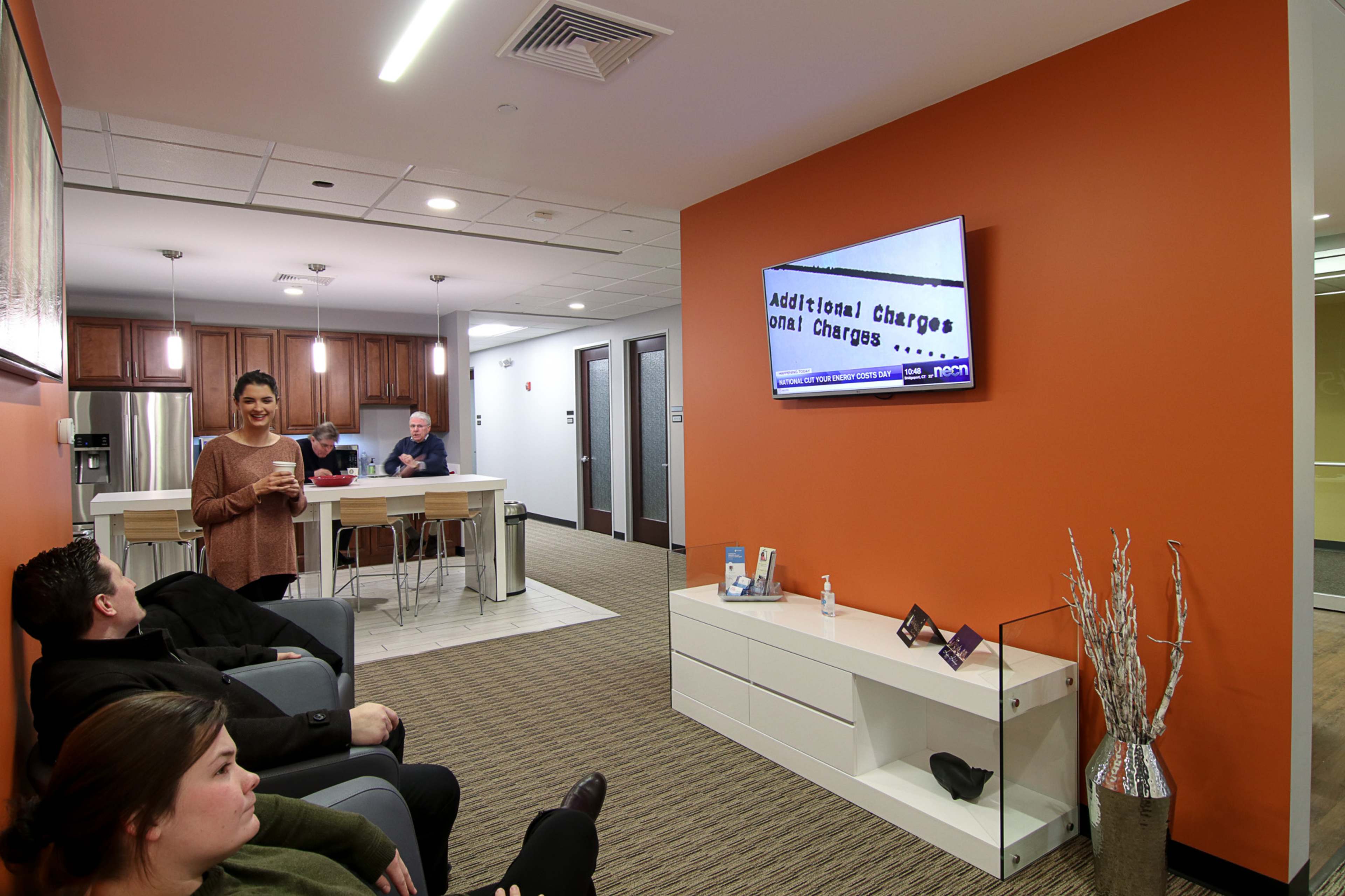 A woman stands in a waiting area with orange walls, while two people sit on a couch and another person works at a kitchen counter in the background.