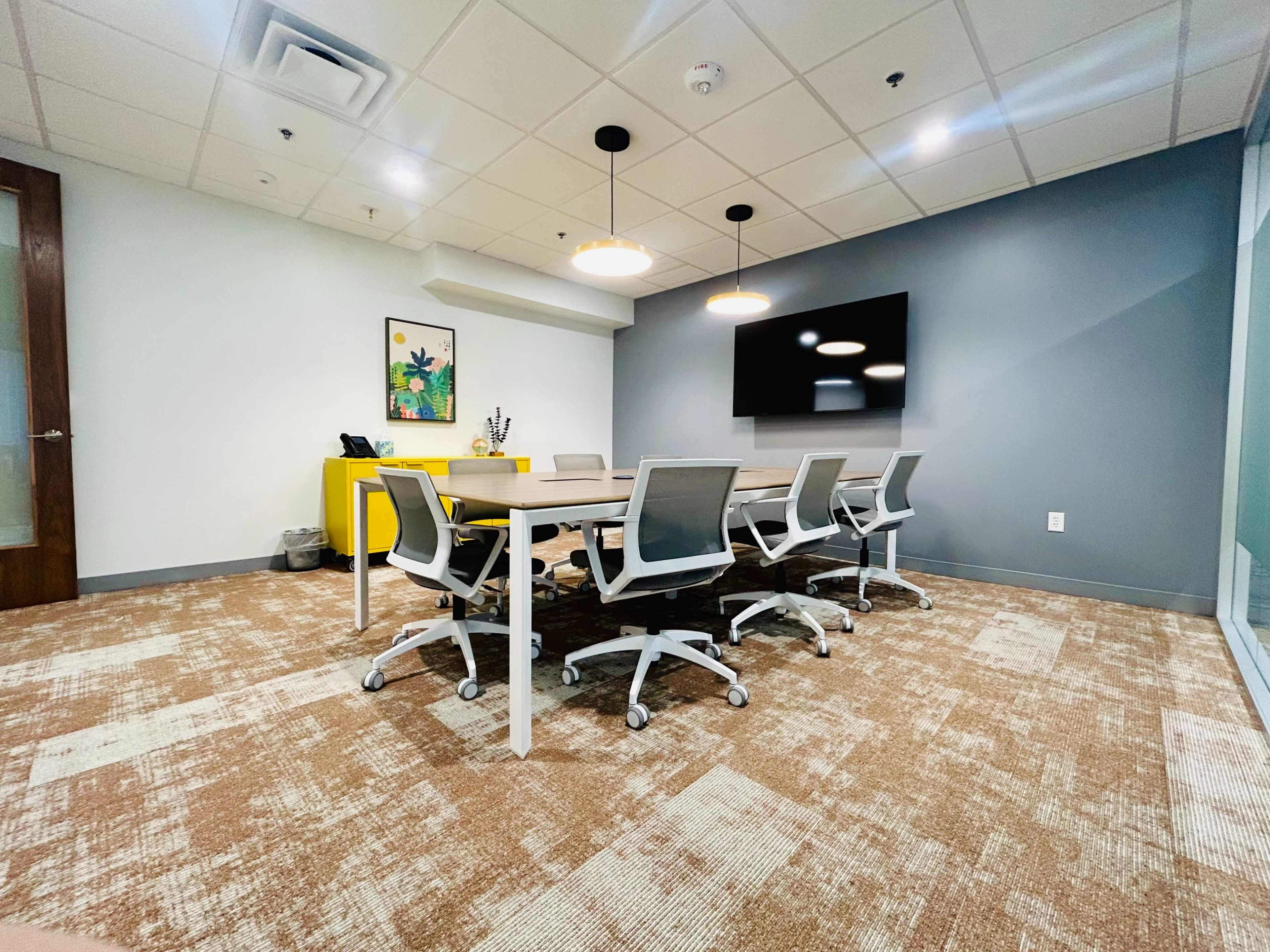 A modern conference room features a large table surrounded by six office chairs, with a television on the wall and a yellow cabinet in the background.