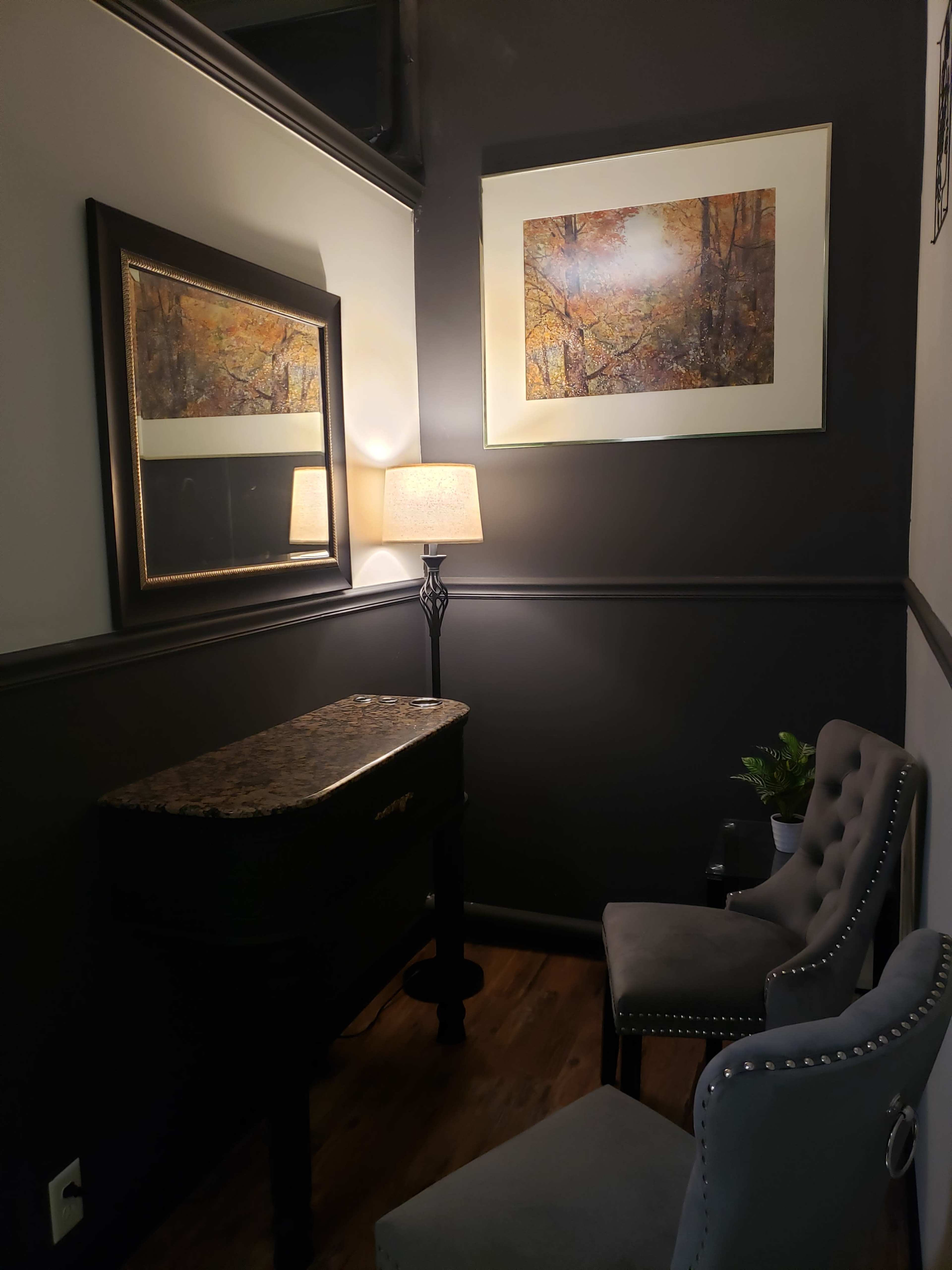 The image shows a corner of a room featuring a dark wall, a framed artwork, a mirror, a lamp, a small table, and two chairs.