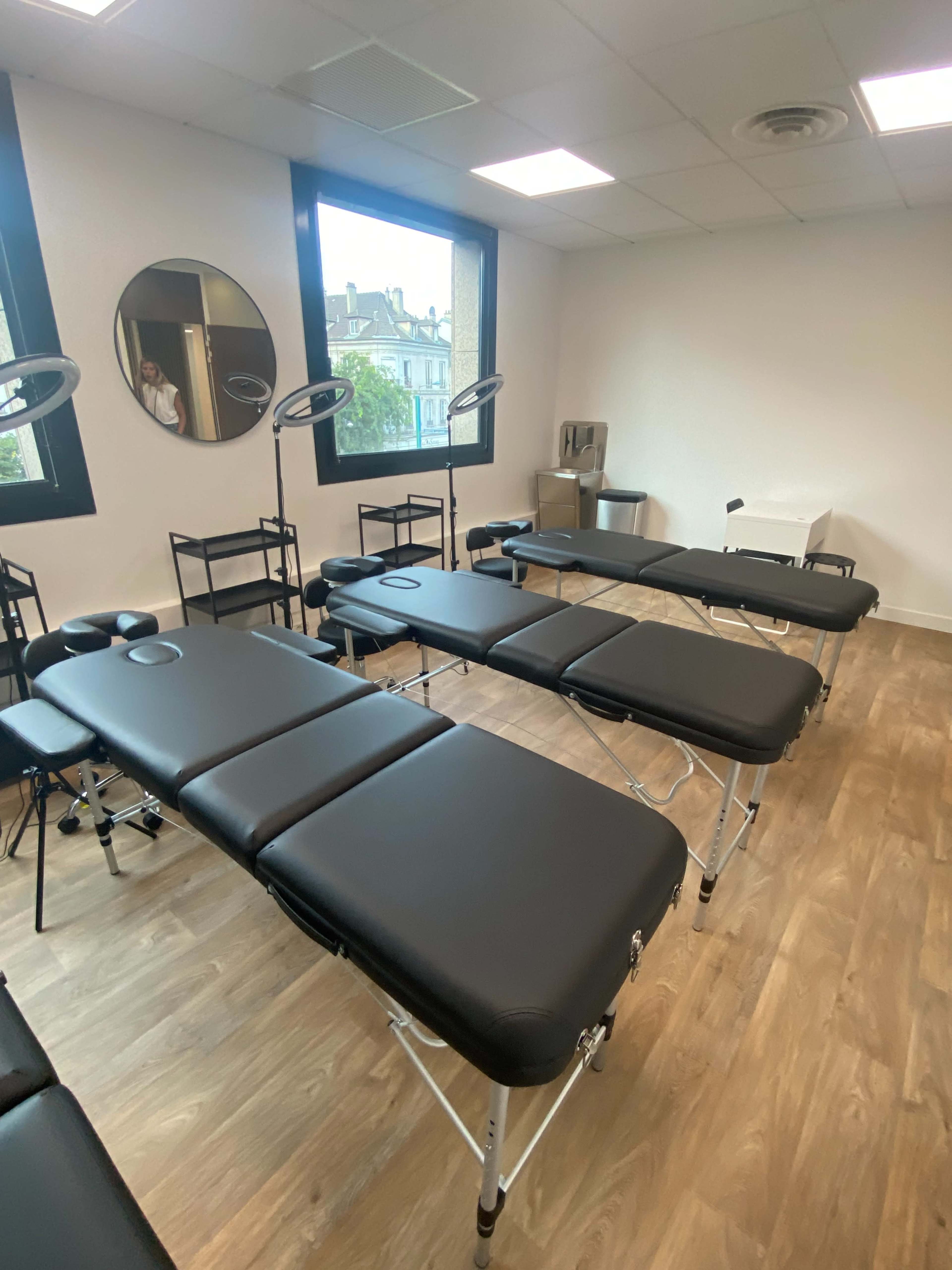 A spacious treatment room featuring several black massage tables arranged in a well-lit area with large windows.