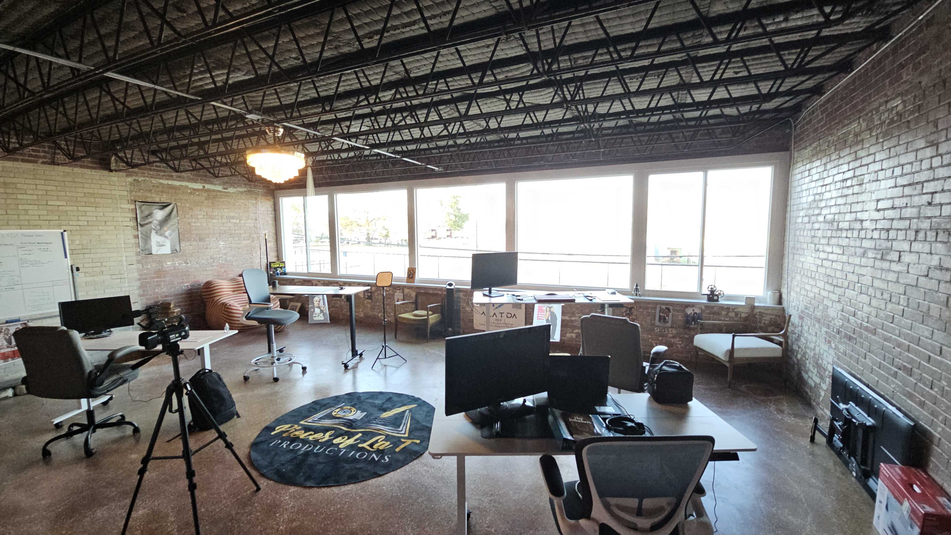 The image shows a spacious office area with multiple desks, chairs, and monitors, illuminated by large windows overlooking an outdoor view.