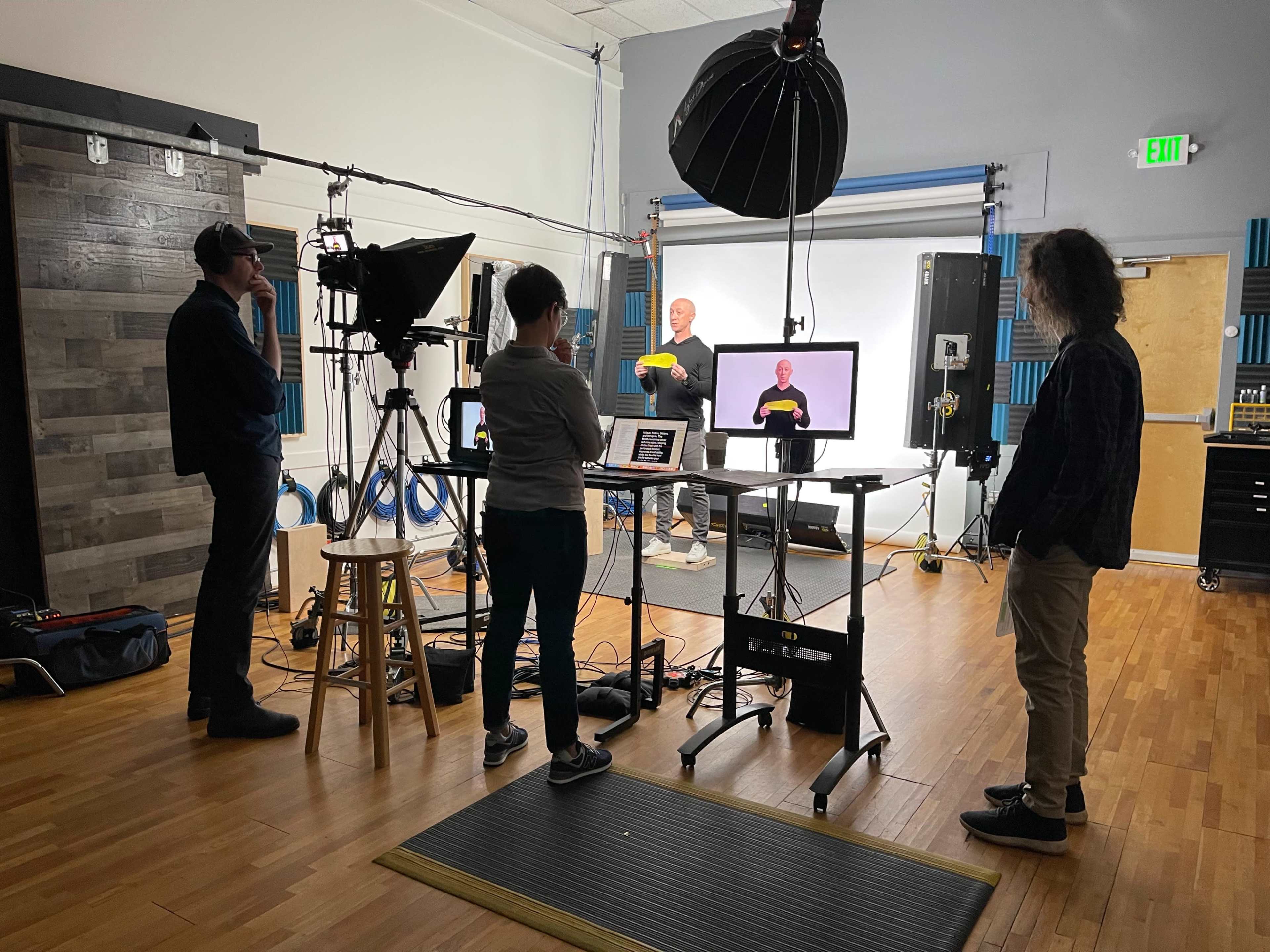 A group of four people is gathered in a studio setting, with one person in front of a screen presenting while the others observe and operate equipment.