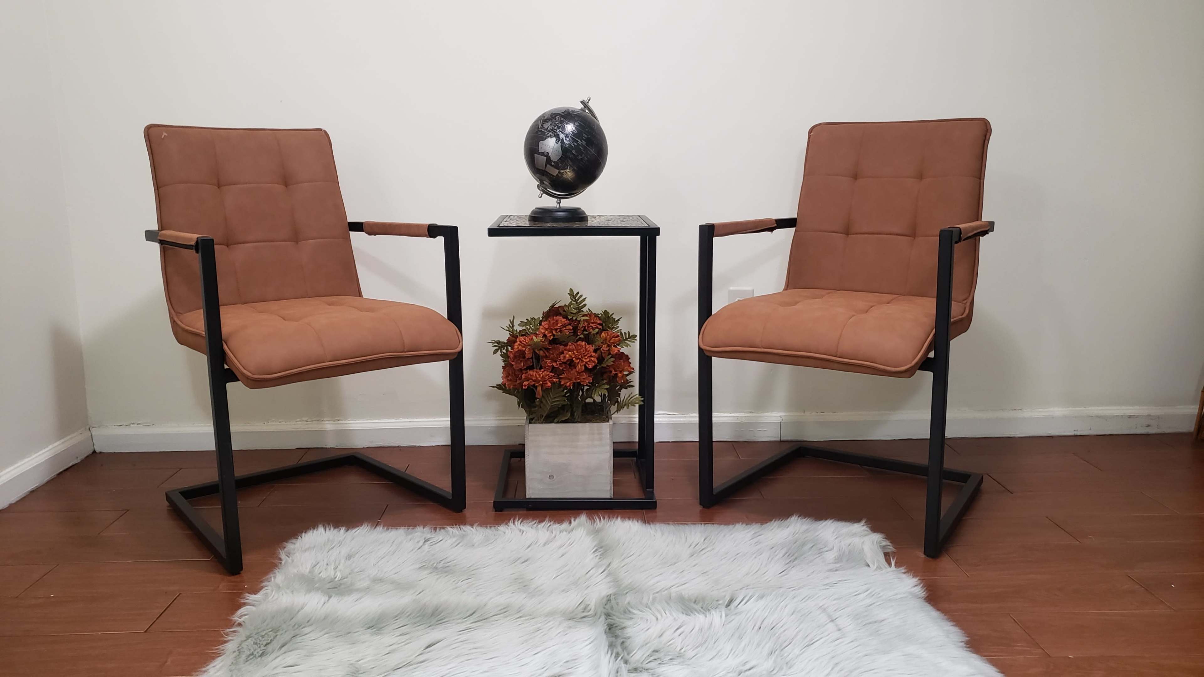 Two brown chairs positioned on either side of a small table with a globe and a floral arrangement, placed on a soft white rug in a room with plain walls.