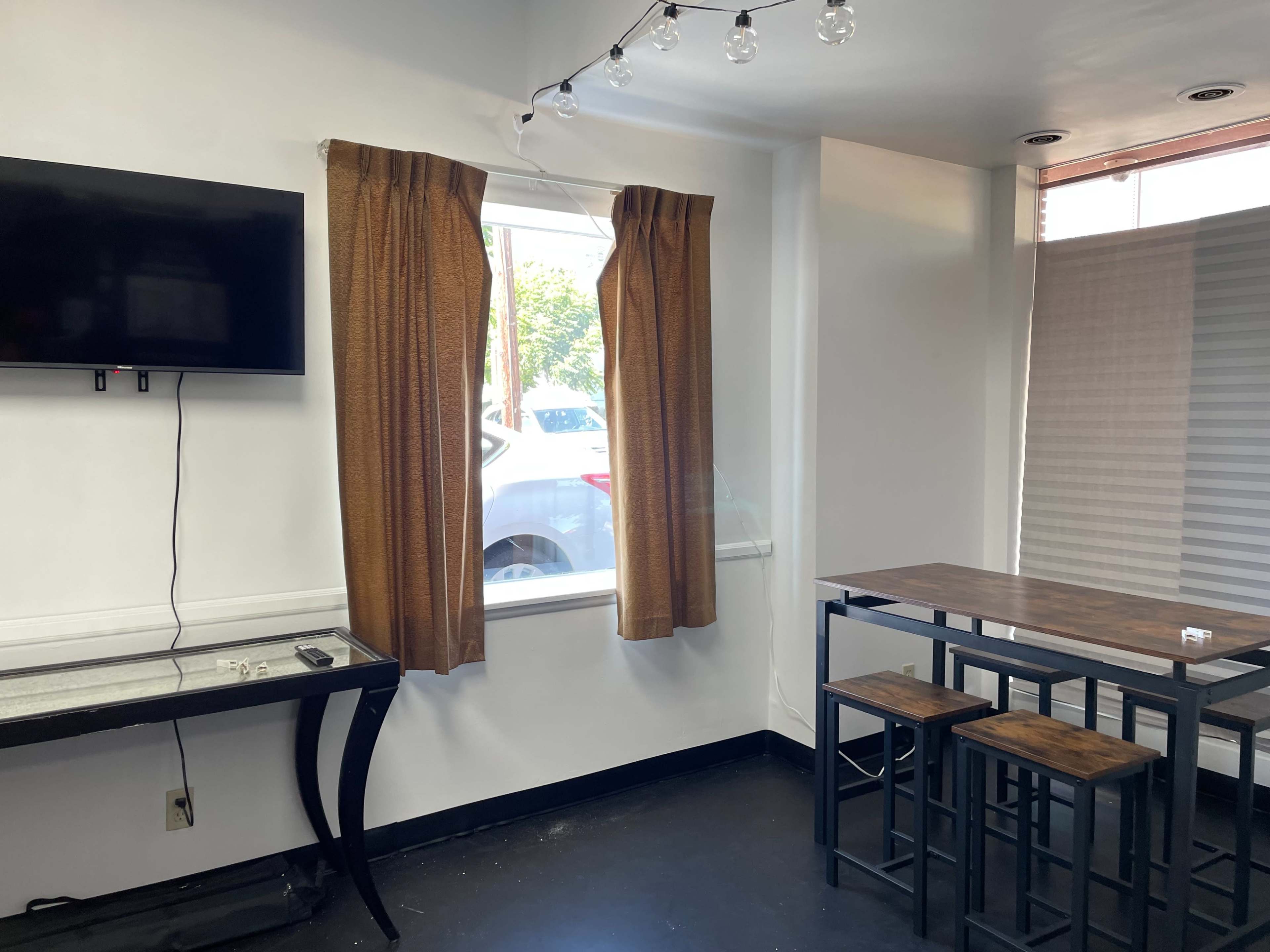 A small room with a television mounted on the wall, a table with stools, and a window covered by brown curtains.