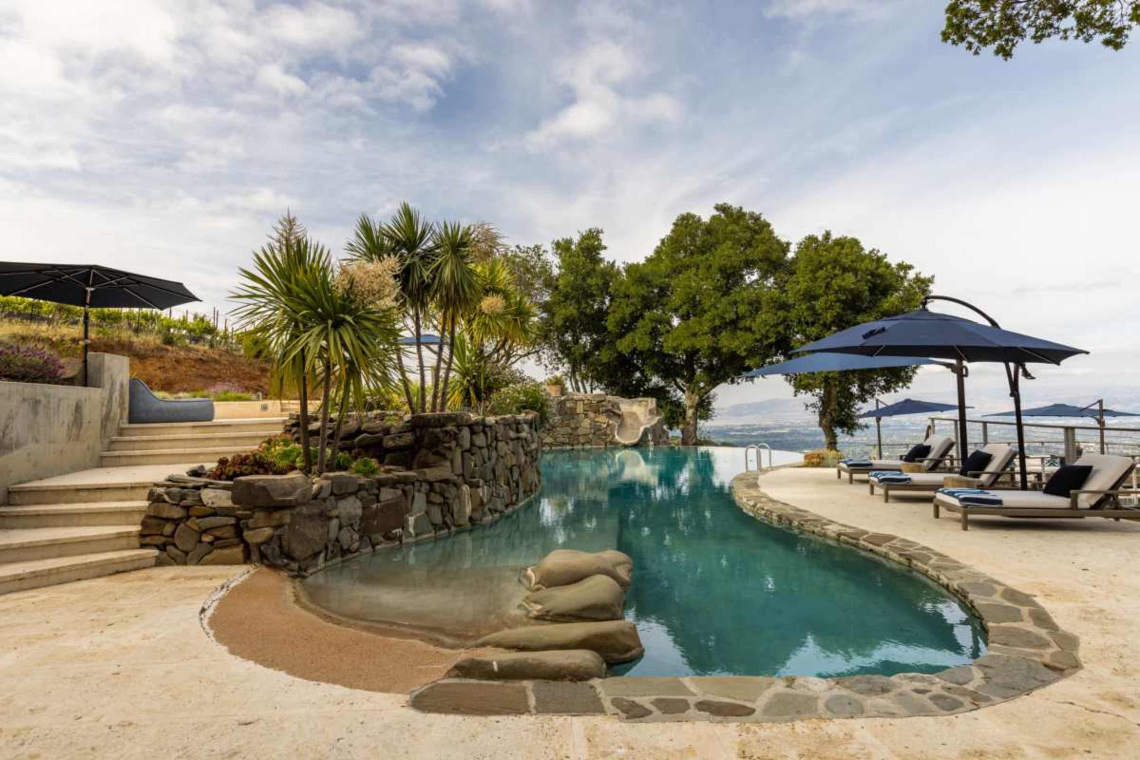 The image shows a curved swimming pool surrounded by rock formations, palm trees, and lounge chairs near a body of water, under a partly cloudy sky.
