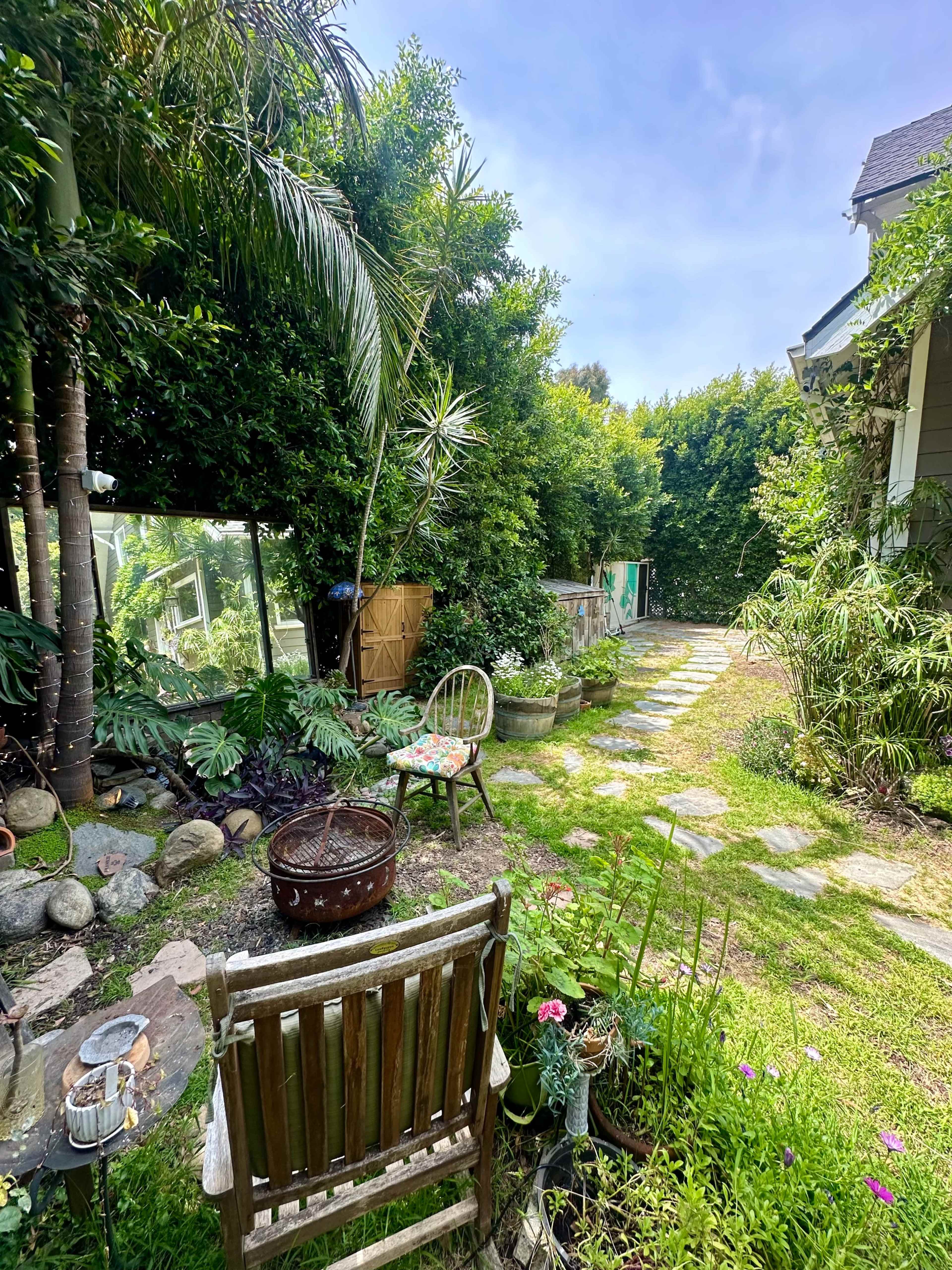 A path lined with stepping stones leads through a garden filled with lush greenery, decorative plants, and a wooden chair.