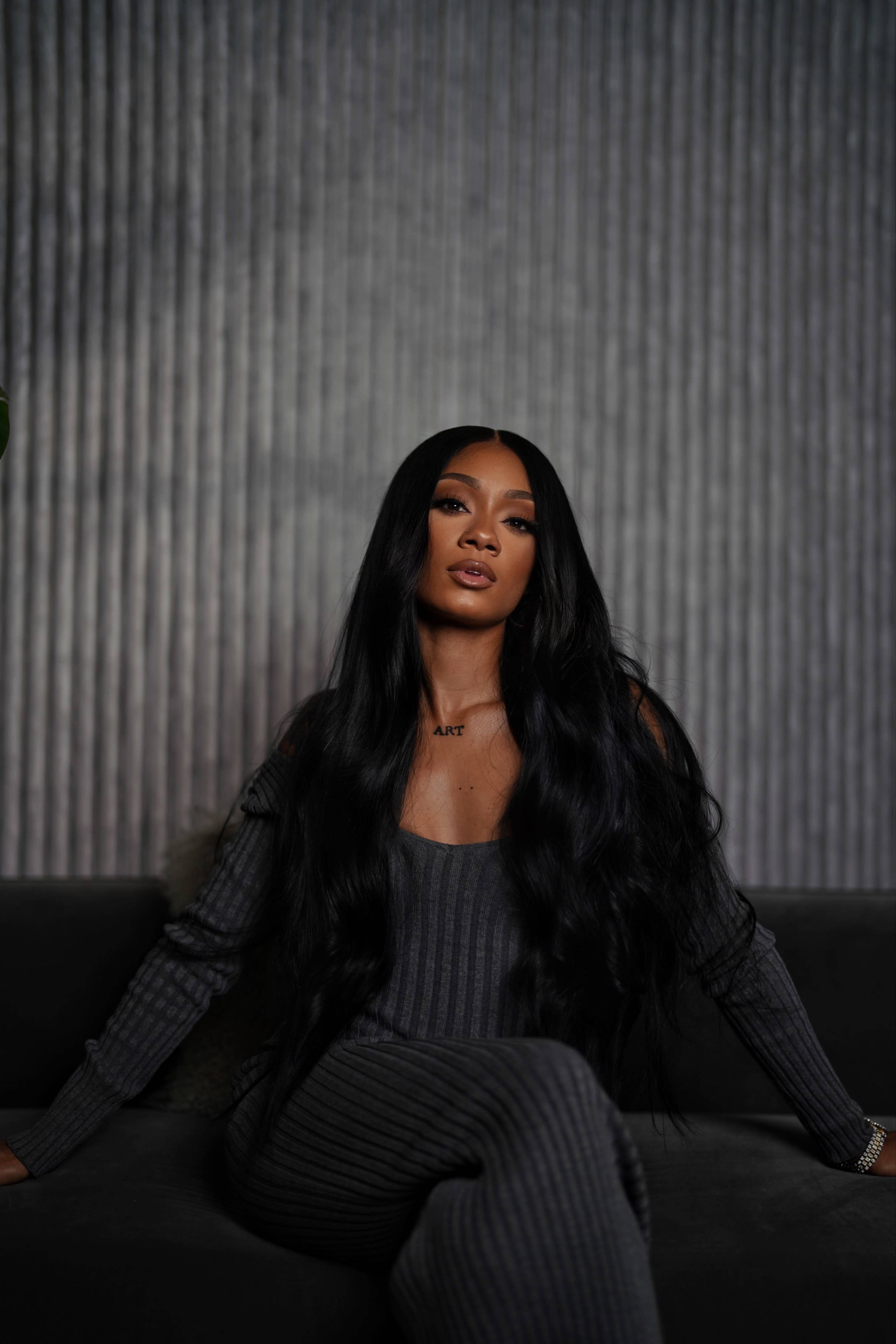 A person with long black hair sits on a dark sofa against a textured gray wall, wearing a form-fitting gray outfit.