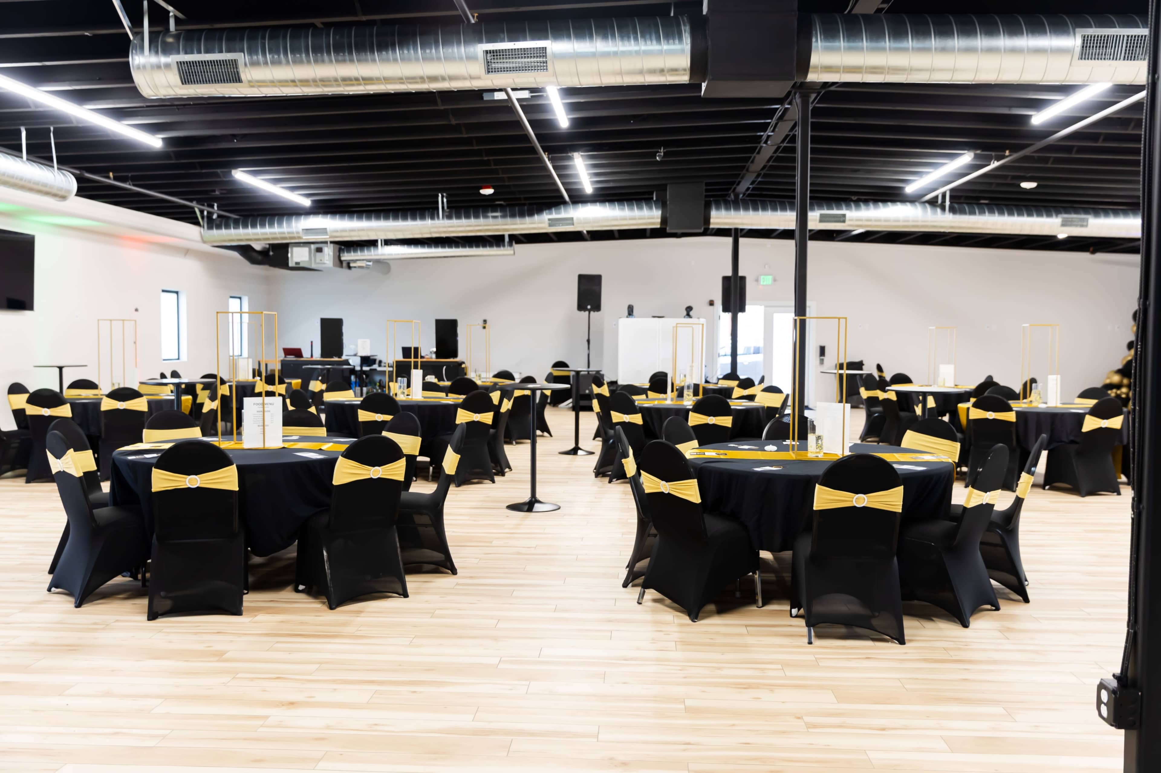 The image shows a spacious event venue with several round tables covered in black tablecloths adorned with gold accents, arranged neatly on a wooden floor.