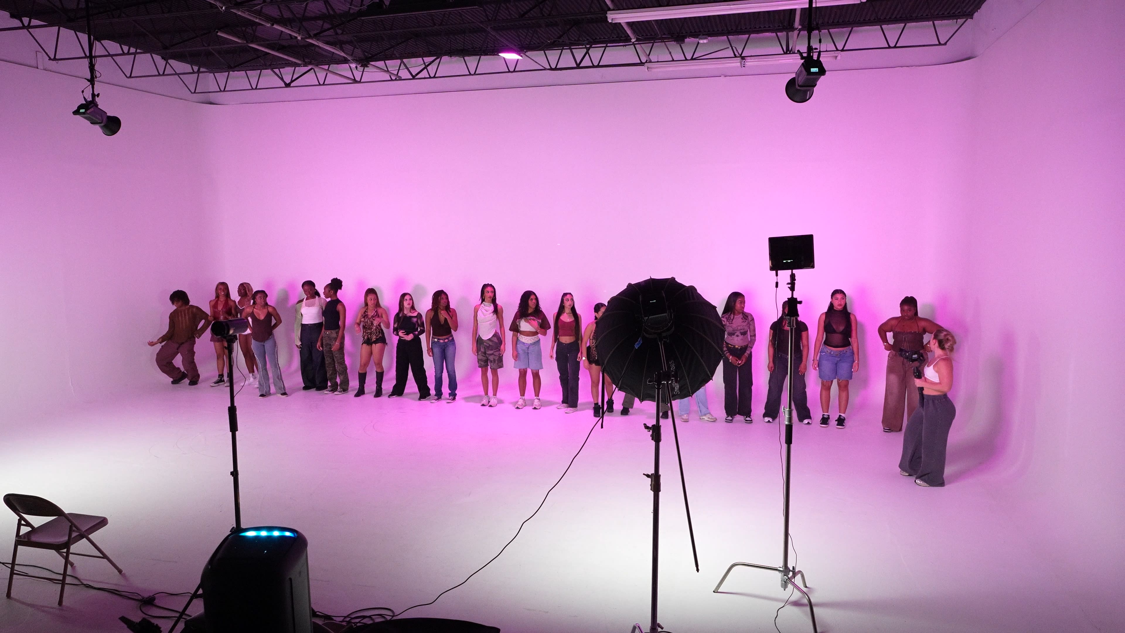 A group of individuals stands in a line on a bright, pink-lit stage set against a plain white backdrop.