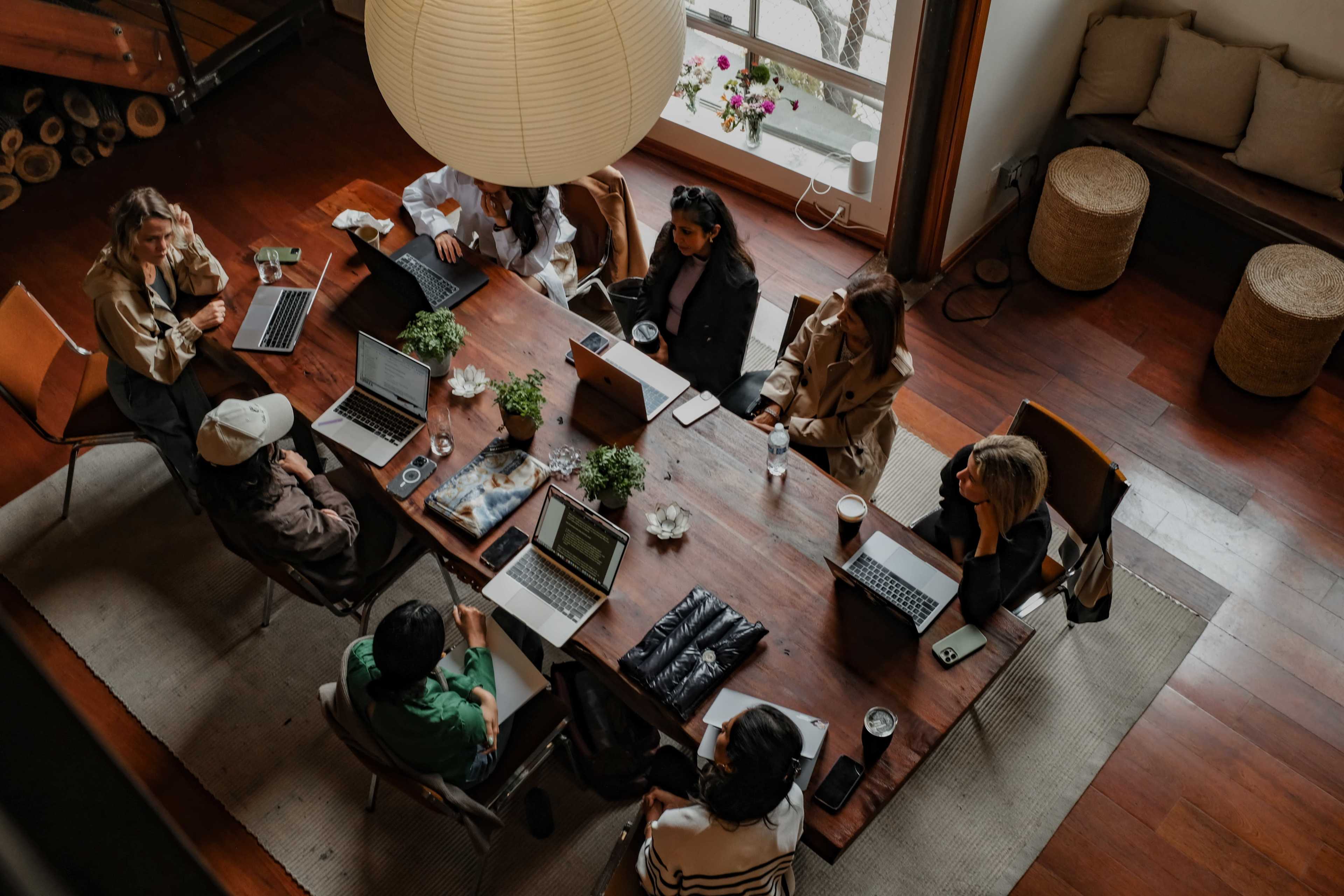 A group of eight individuals is seated around a large wooden table in a well-lit room, each working on laptops and engaging in conversation.