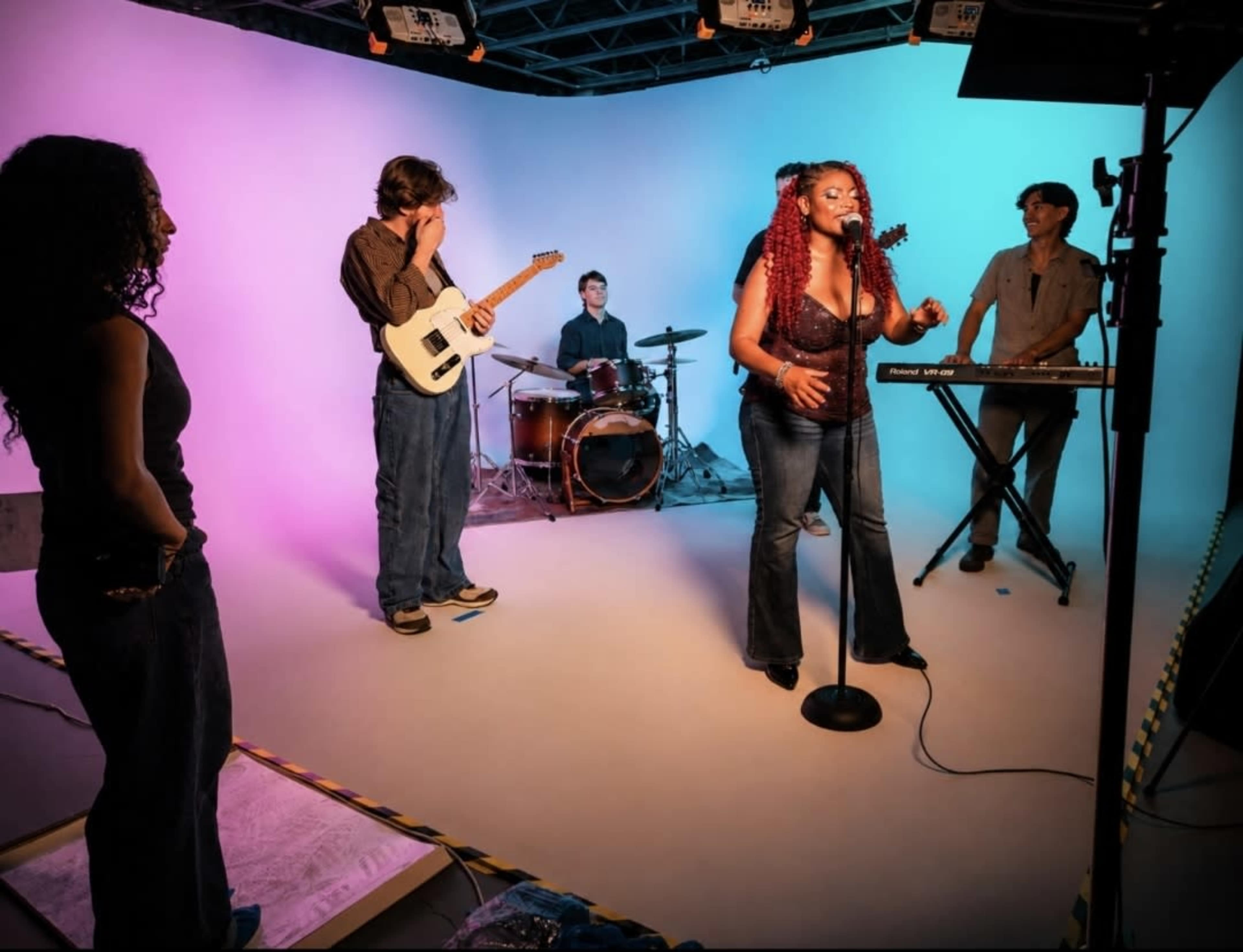 A group of musicians performs on a brightly lit stage with a colorful backdrop, featuring a vocalist, guitarist, drummer, and keyboardist.