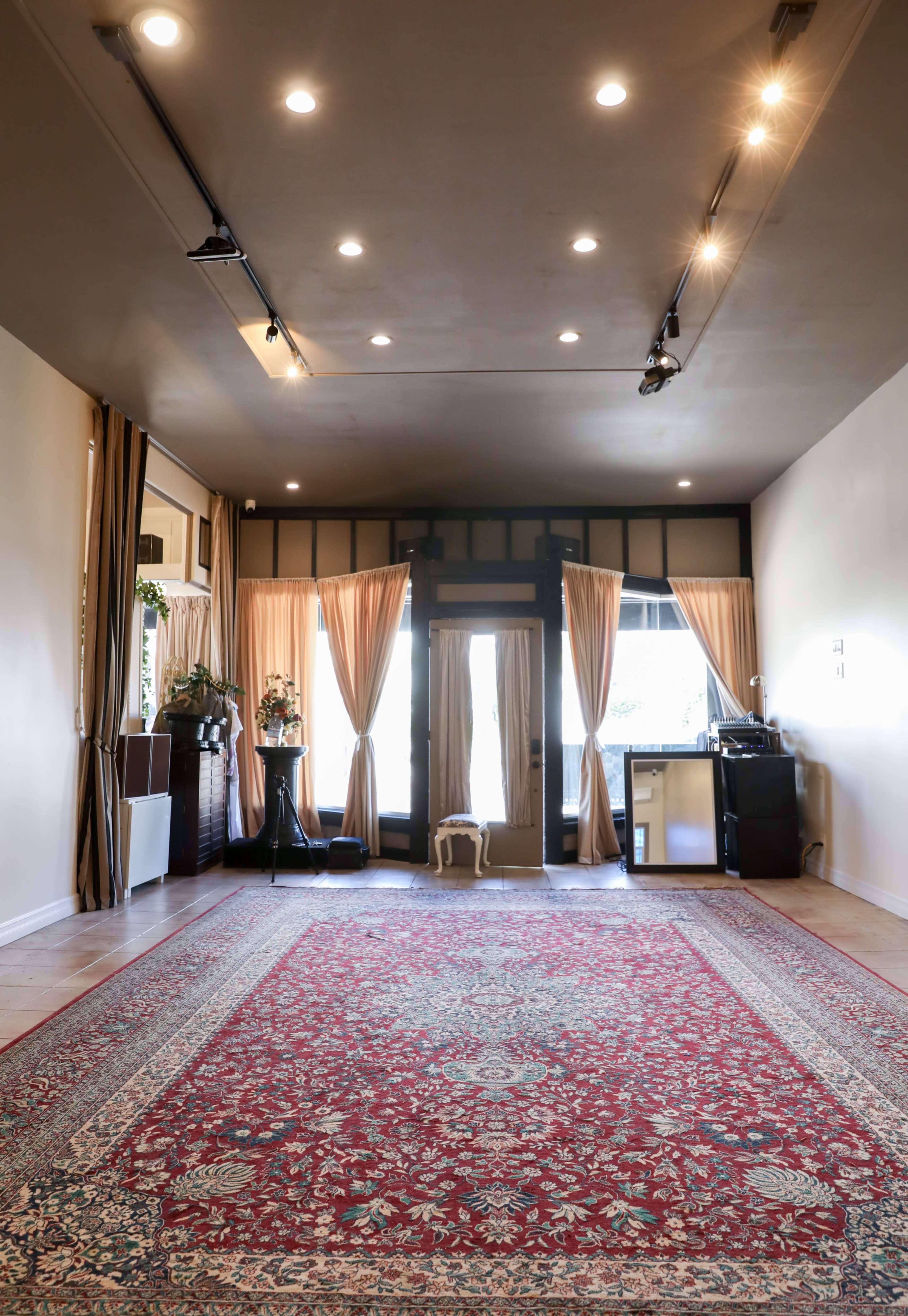 A spacious room with hardwood floors, large windows adorned with beige curtains, a decorative area rug, and minimal furniture.