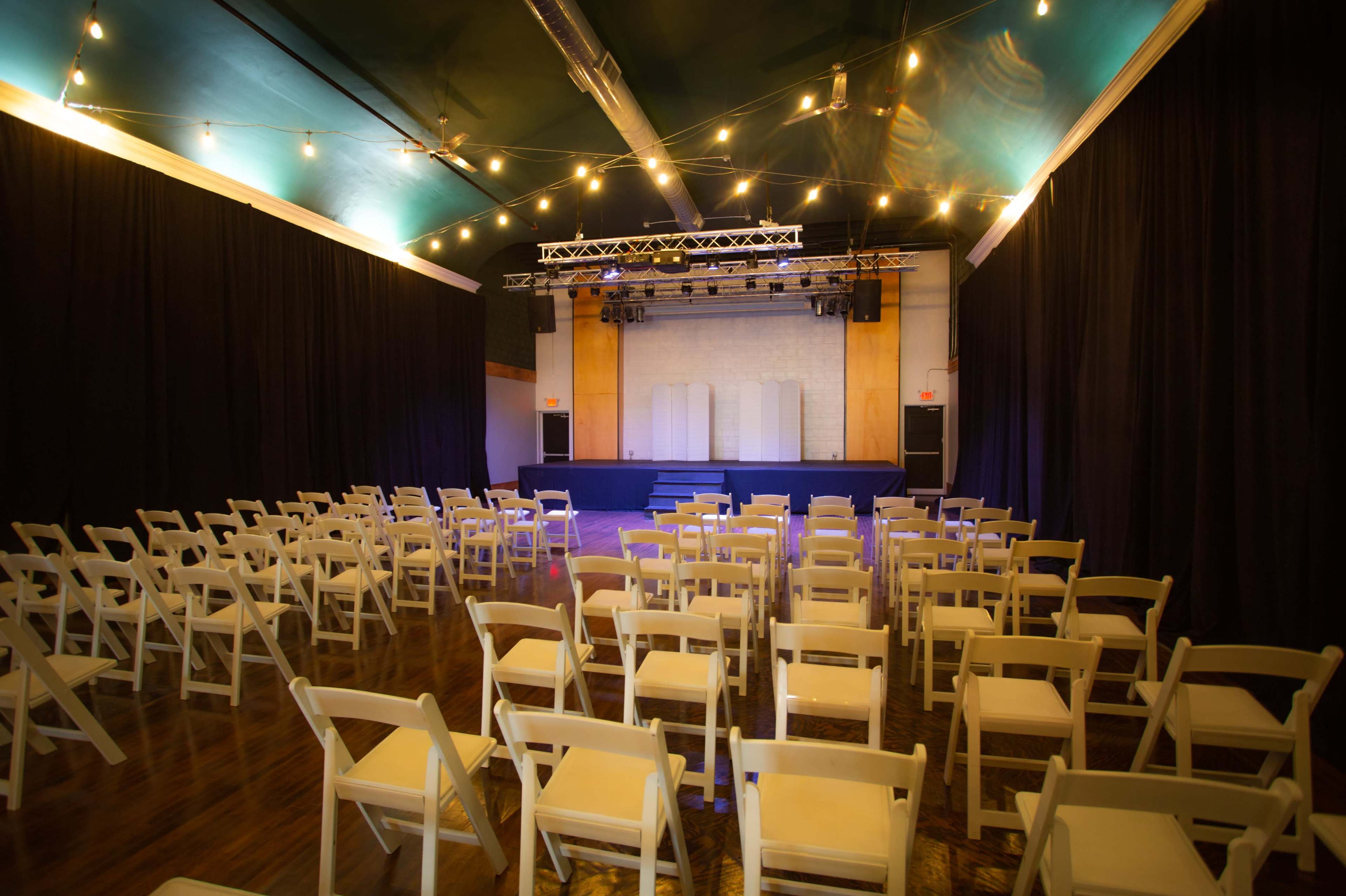 The image shows a stage set up in a venue with rows of empty white folding chairs arranged in front of it, illuminated by overhead string lights.